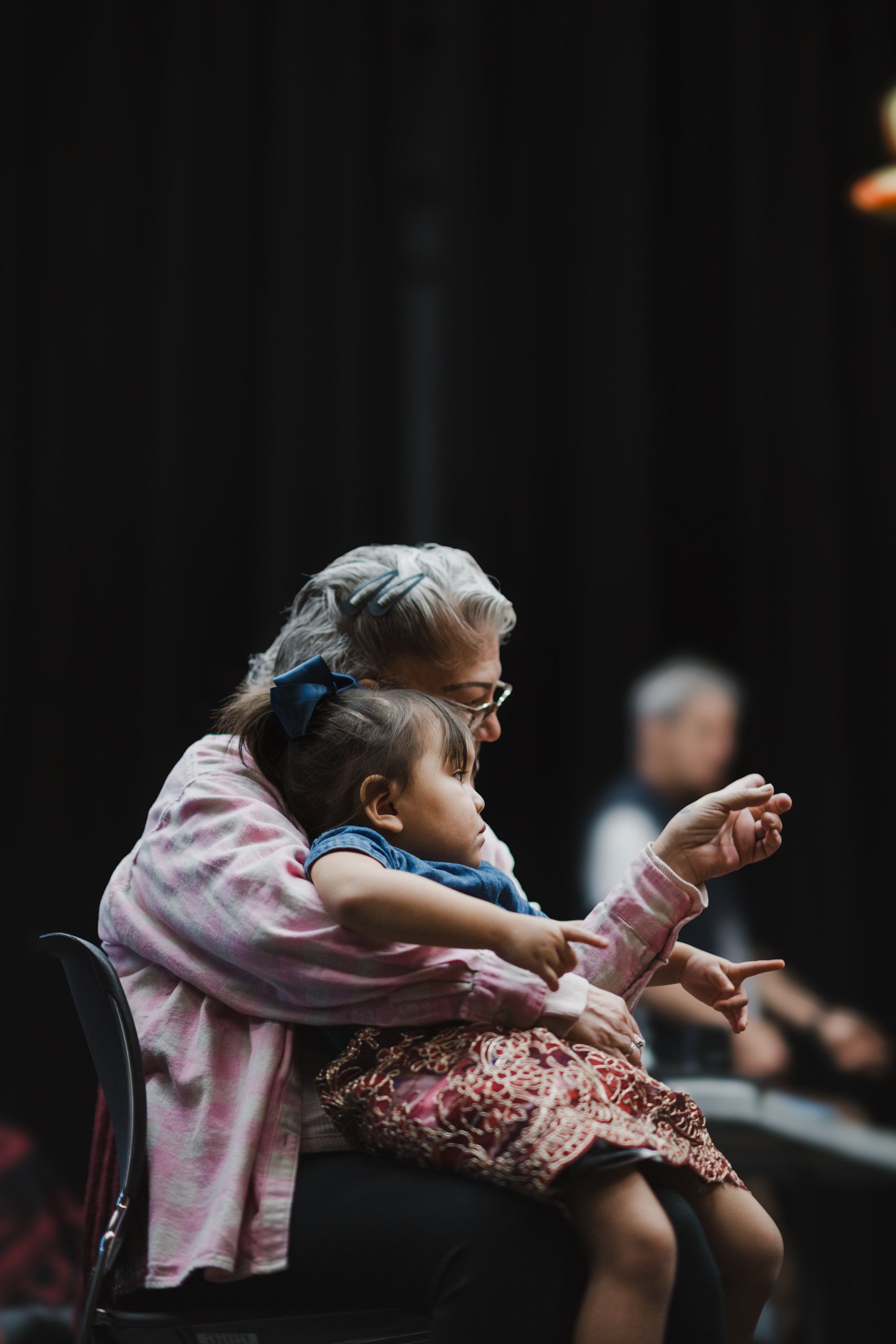 Grandmother with silver hair and glasses holds a young child pointing outward during a Coast Salish performance, dark curtains behind them