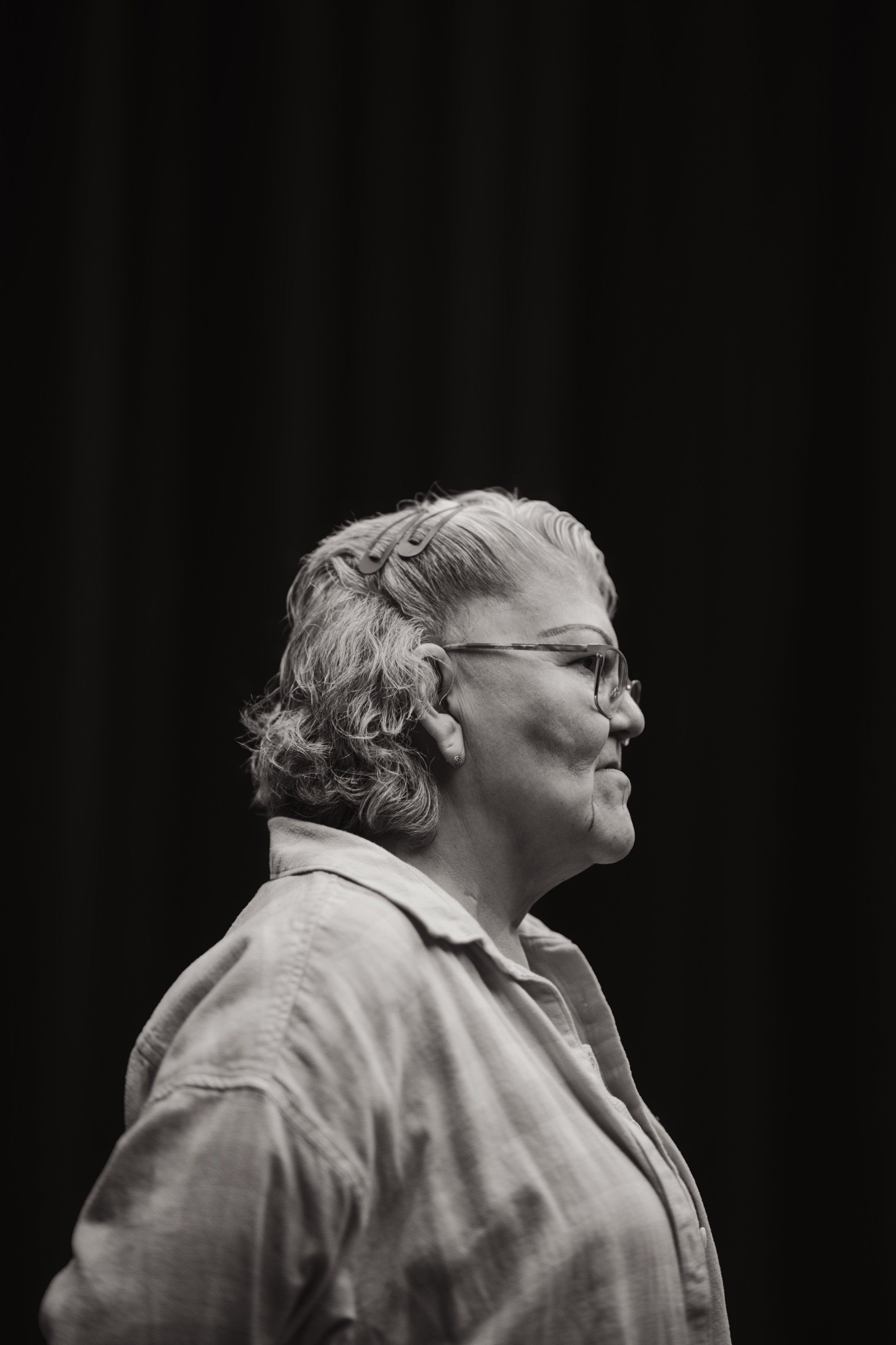 Profile portrait of an older man with grey wavy hair and glasses, wearing a light-coloured shirt against a dark background