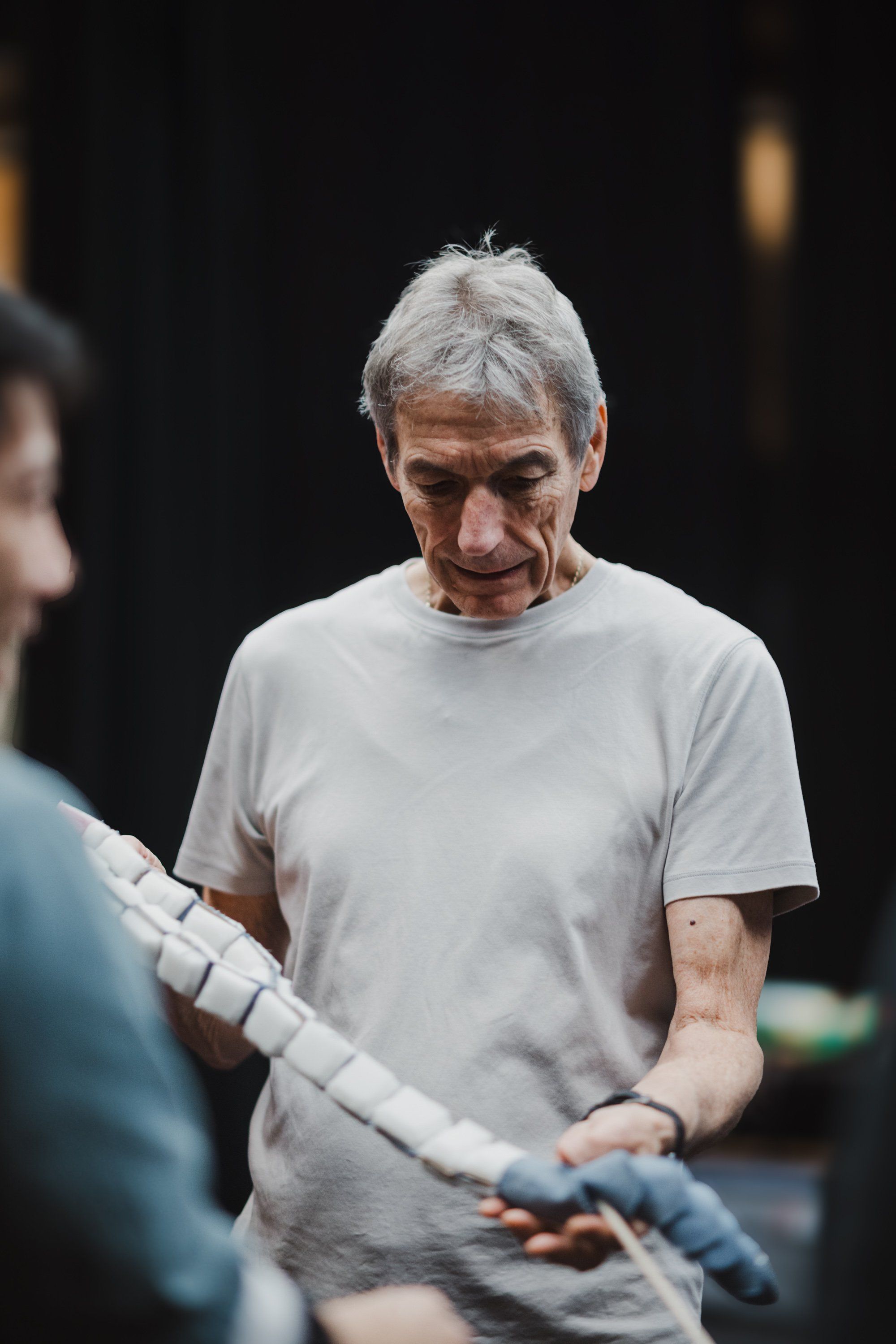 An elder with white hair in a light gray t-shirt holds bundled white fabric, looking down with gentle focus during a Coast Salish cultural celebration