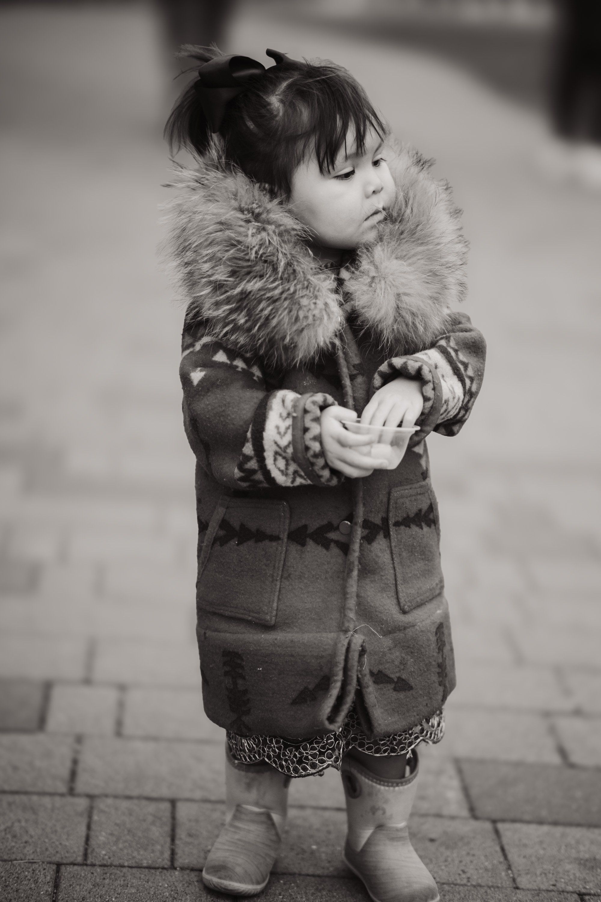 Young child in fur-trimmed winter coat and patterned mittens stands on a brick pathway in black and white portrait