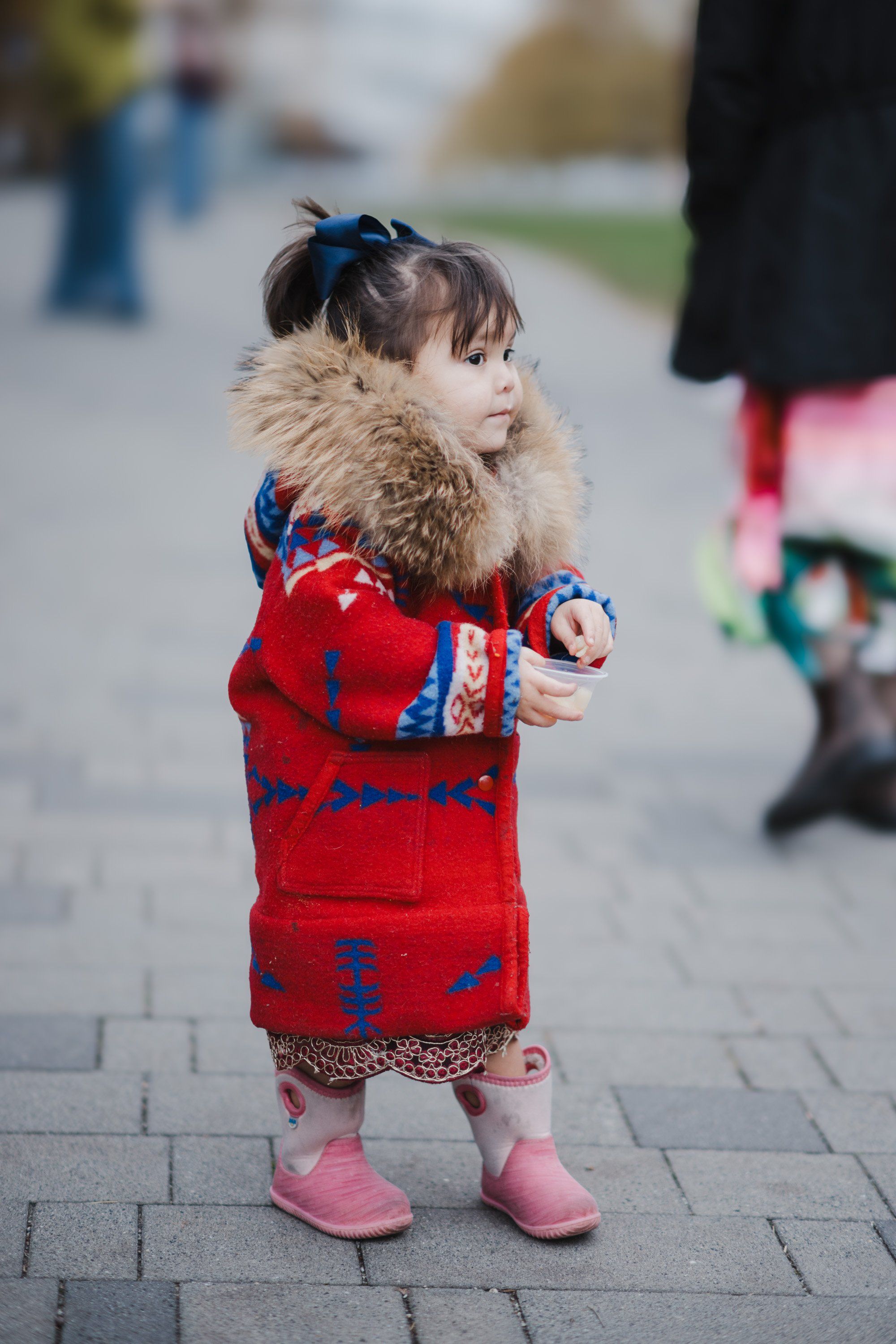 Toddler in red patterned sweater with fur collar stands on brick pavement, gazing thoughtfully to the side