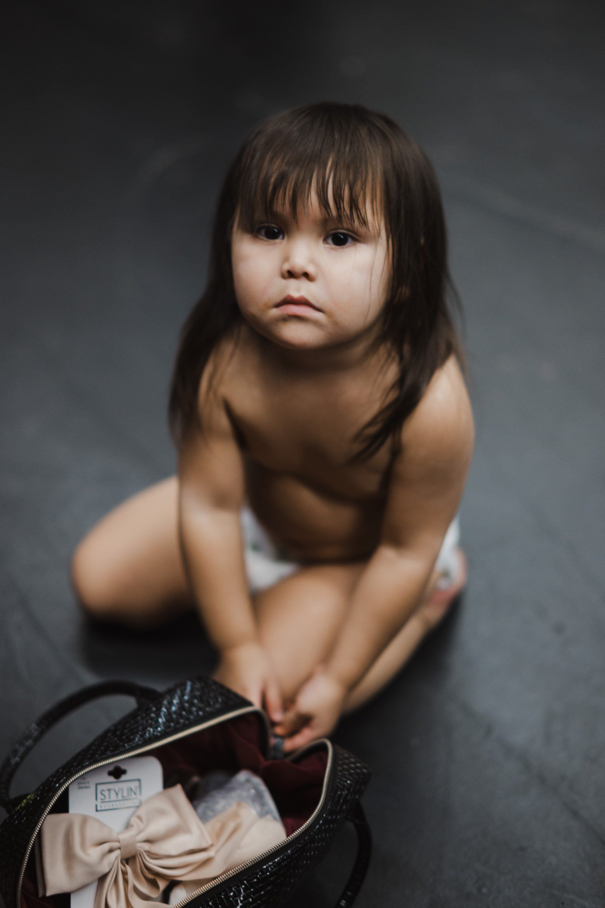 Young child sitting barefoot on gray floor in contemplative pose, holding a woven basket, soft directional lighting