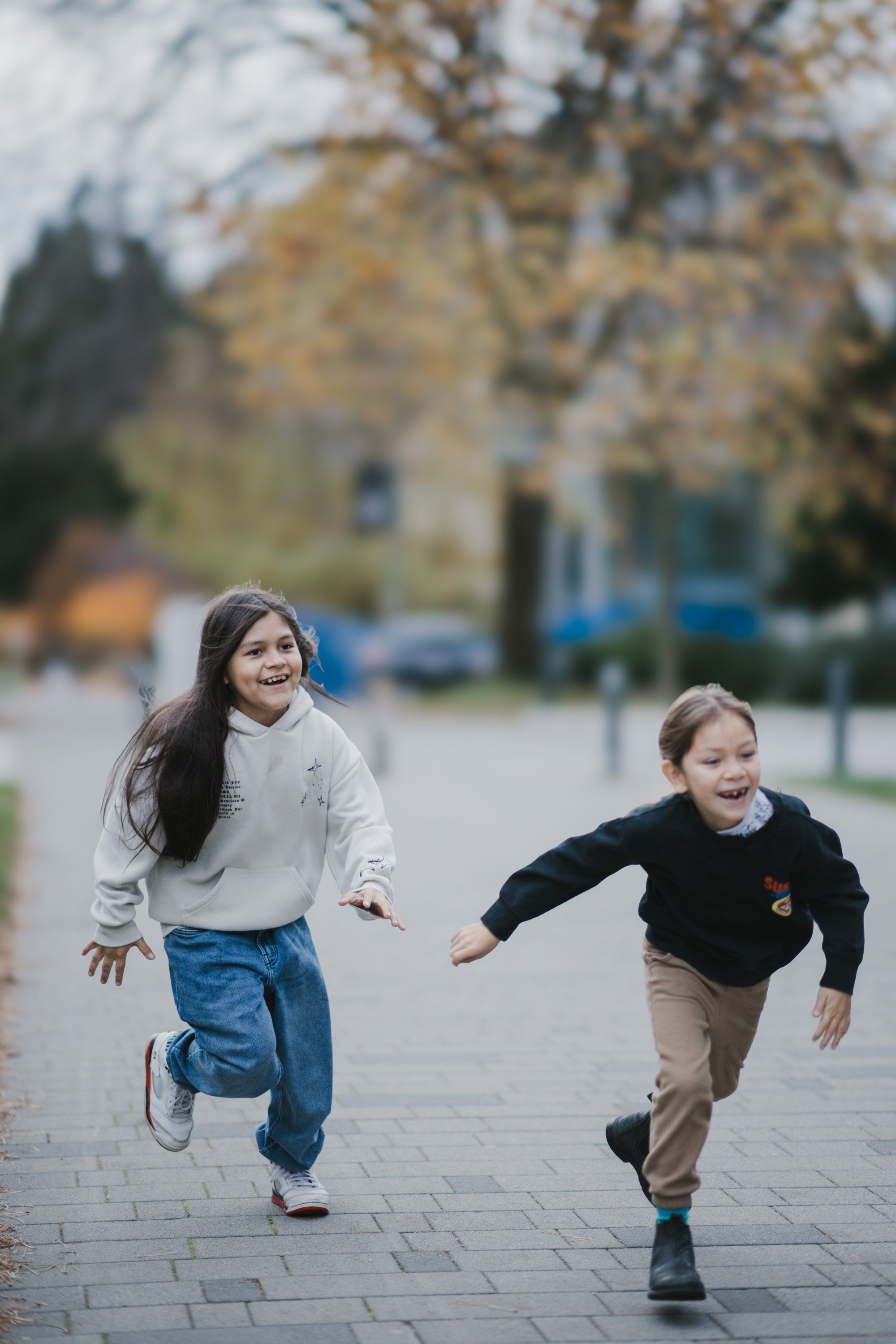 Two children run joyfully across a brick plaza with autumn trees and a stone building in soft, overcast light