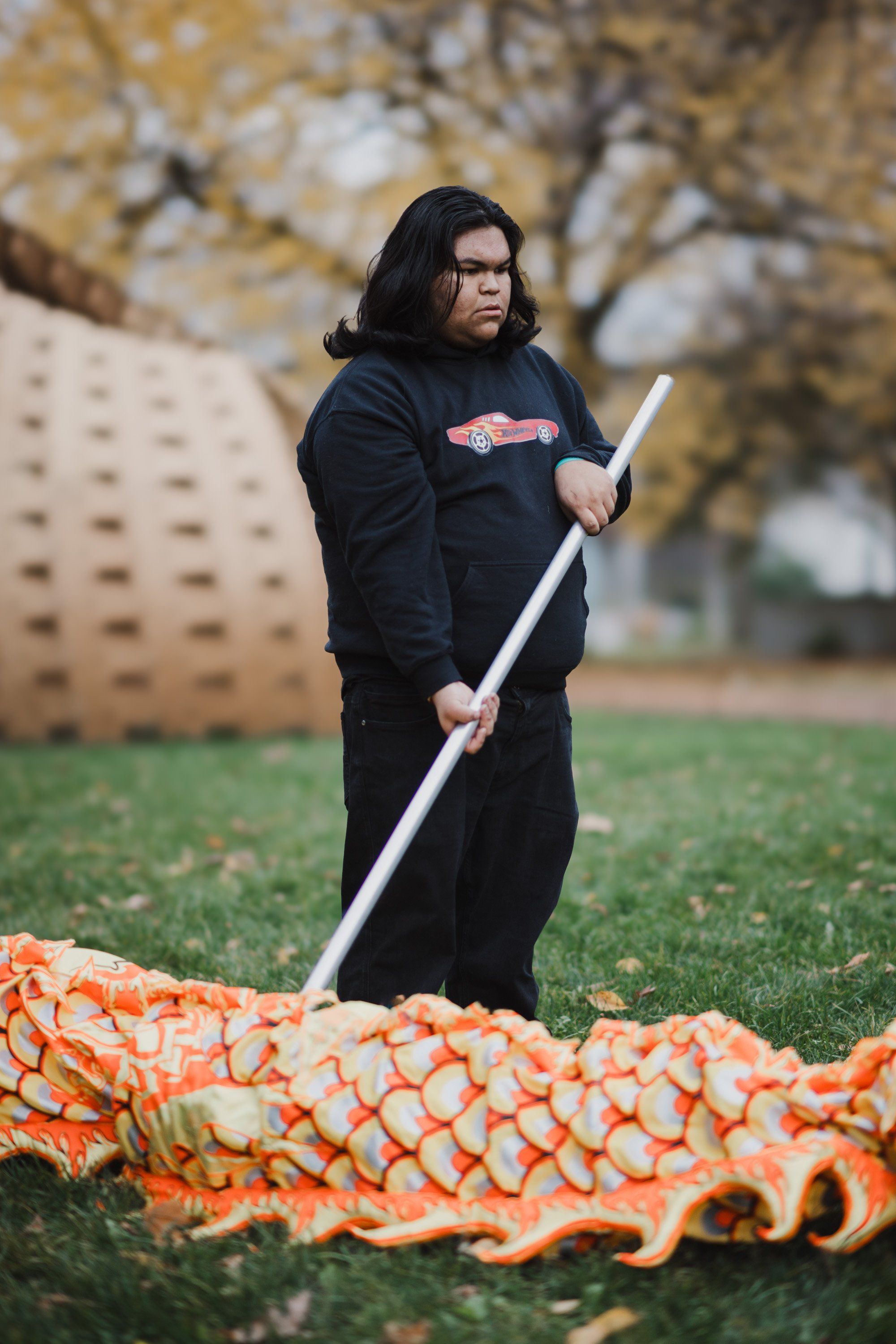 Person in black hoodie holding a carved wooden staff beside an orange and white woven textile on grass, bare trees behind