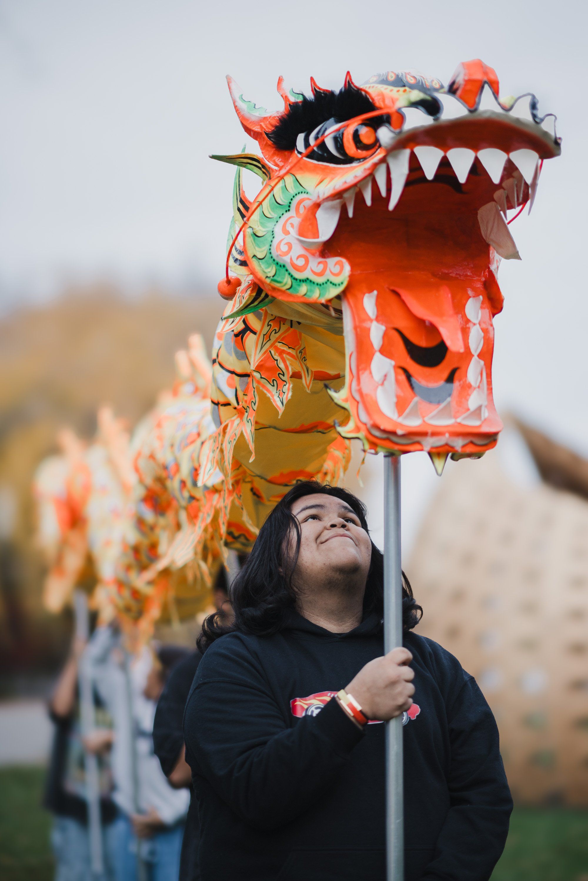Young person in dark clothing gazes upward while holding a vibrant red and gold dragon puppet aloft, autumn trees soft-focused behind them