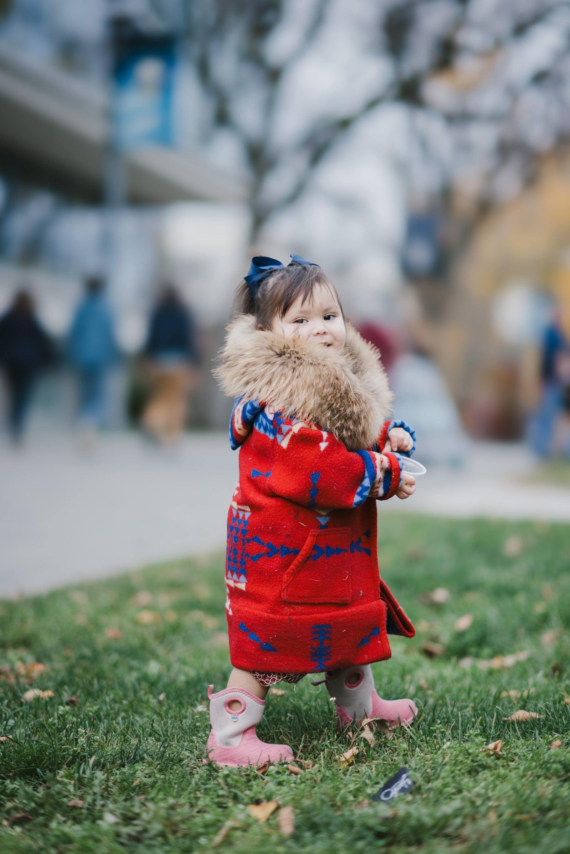 Toddler in red plaid coat with fur collar stands on grass, soft focus architecture behind, gentle afternoon light