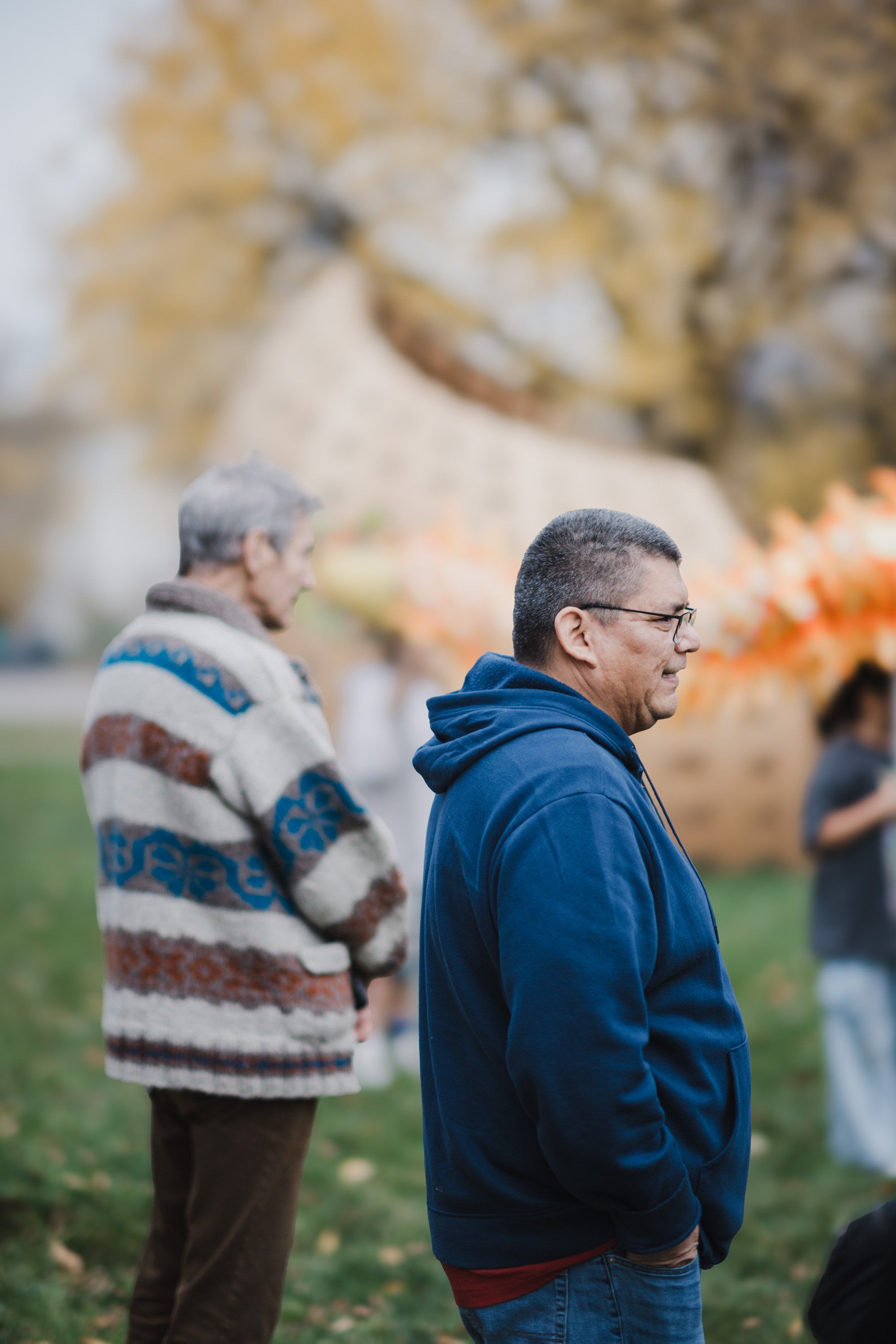 Man in blue hoodie and glasses stands in profile at outdoor gathering, autumn foliage soft behind him