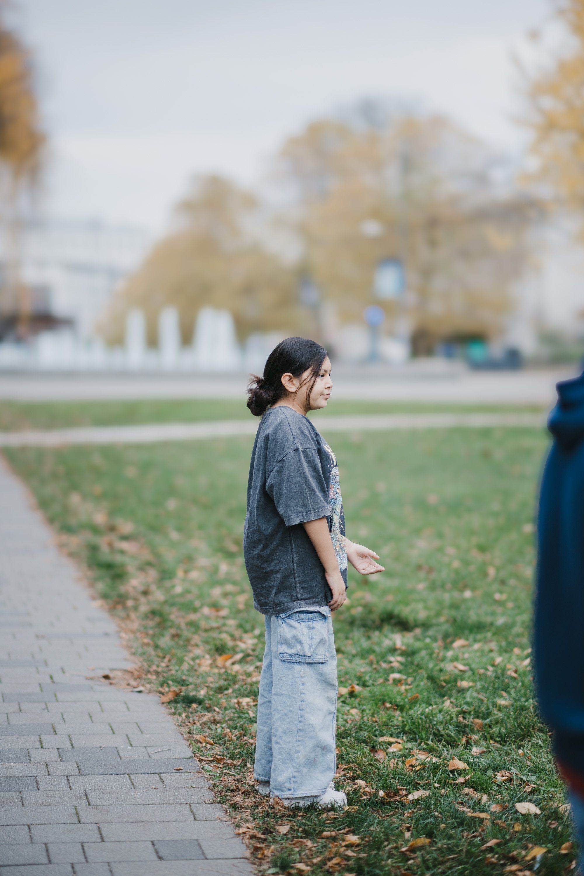 Young person in denim shirt and light skirt standing on a brick pathway beside a green field, autumn trees softly blurred in background
