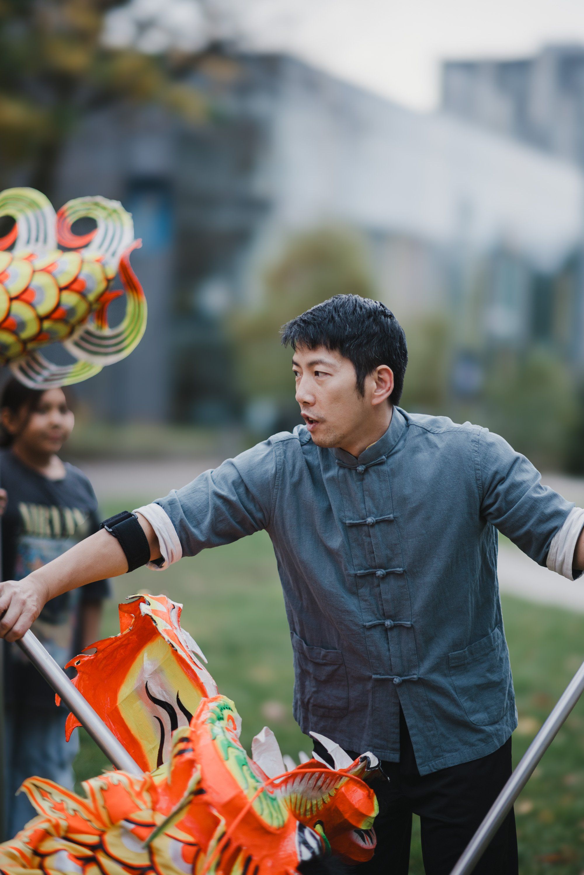 Young man in blue shirt holds traditional Coast Salish kite outdoors with blurred buildings behind, arms outstretched in celebration