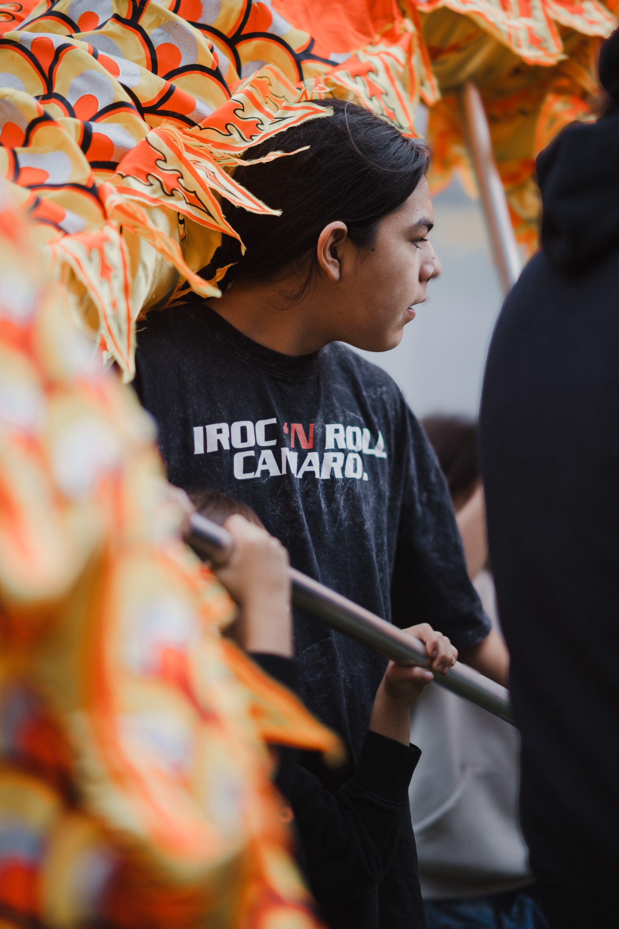 Young person in profile wearing a dark shirt, holding a wooden staff, positioned near vibrant orange and red woven regalia during a Coast Salish cultural cel…