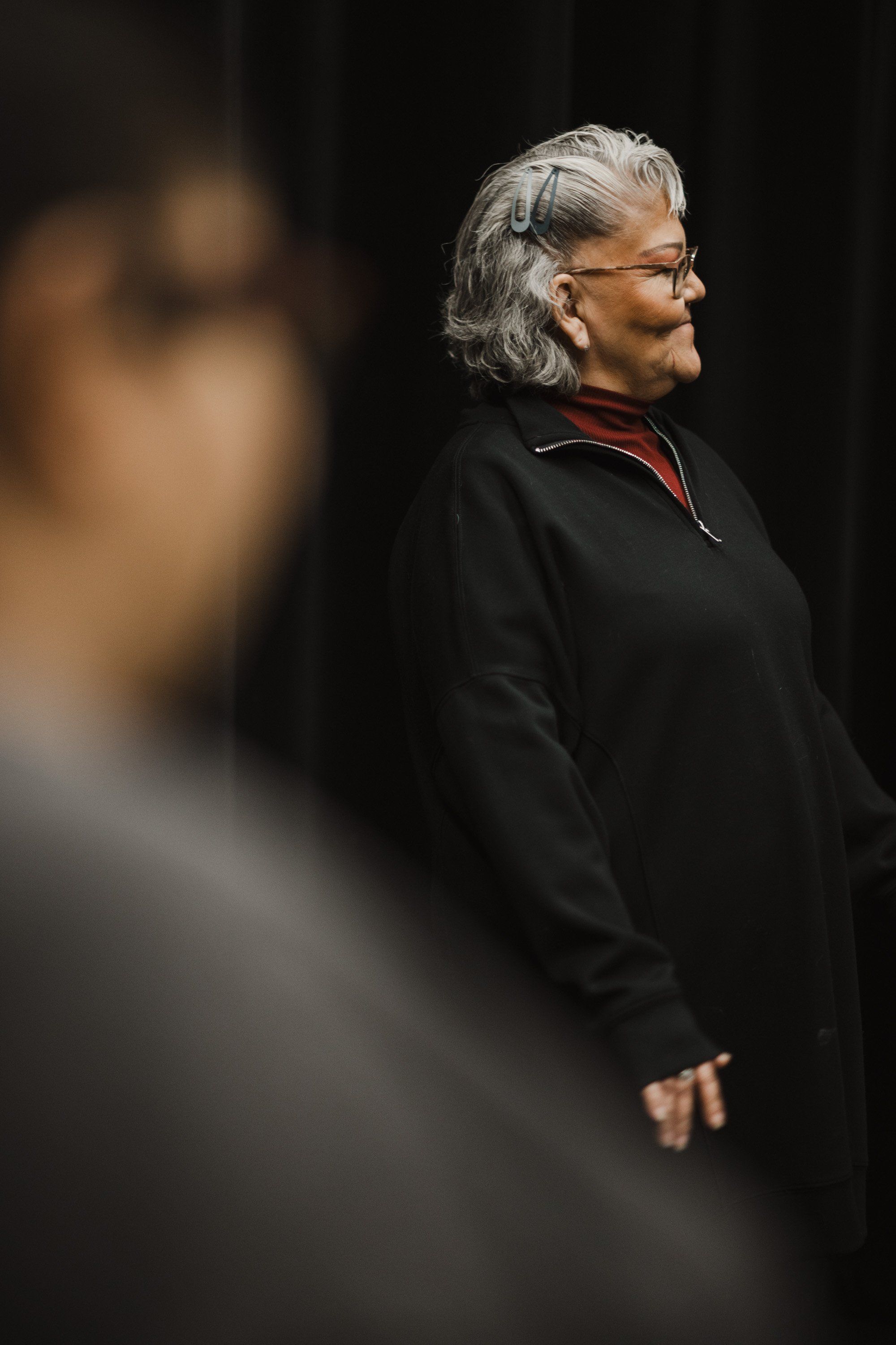 Older woman with silver hair and glasses in profile, wearing black jacket with red collar detail, standing against dark background with soft stage lighting