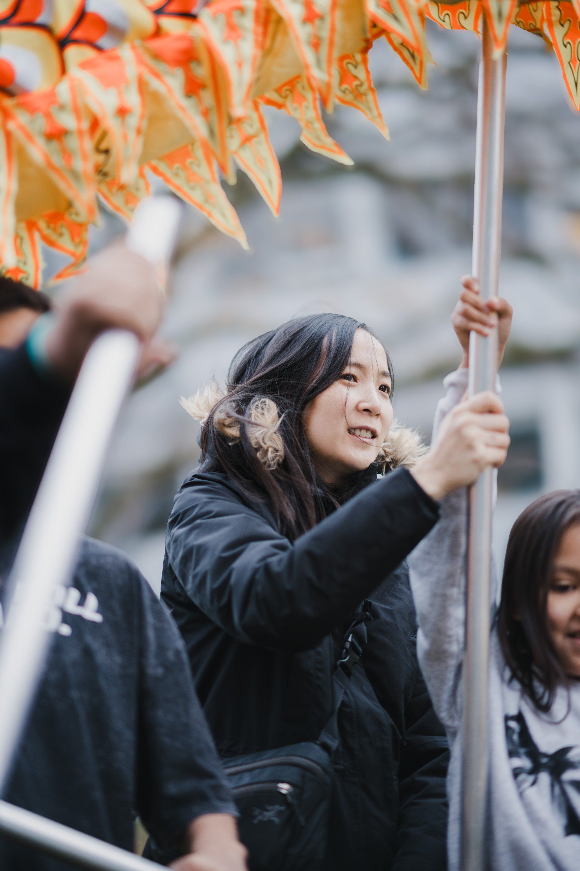 Young woman in black jacket gazes upward while holding a pole beneath golden autumn leaves, smiling during an outdoor celebration