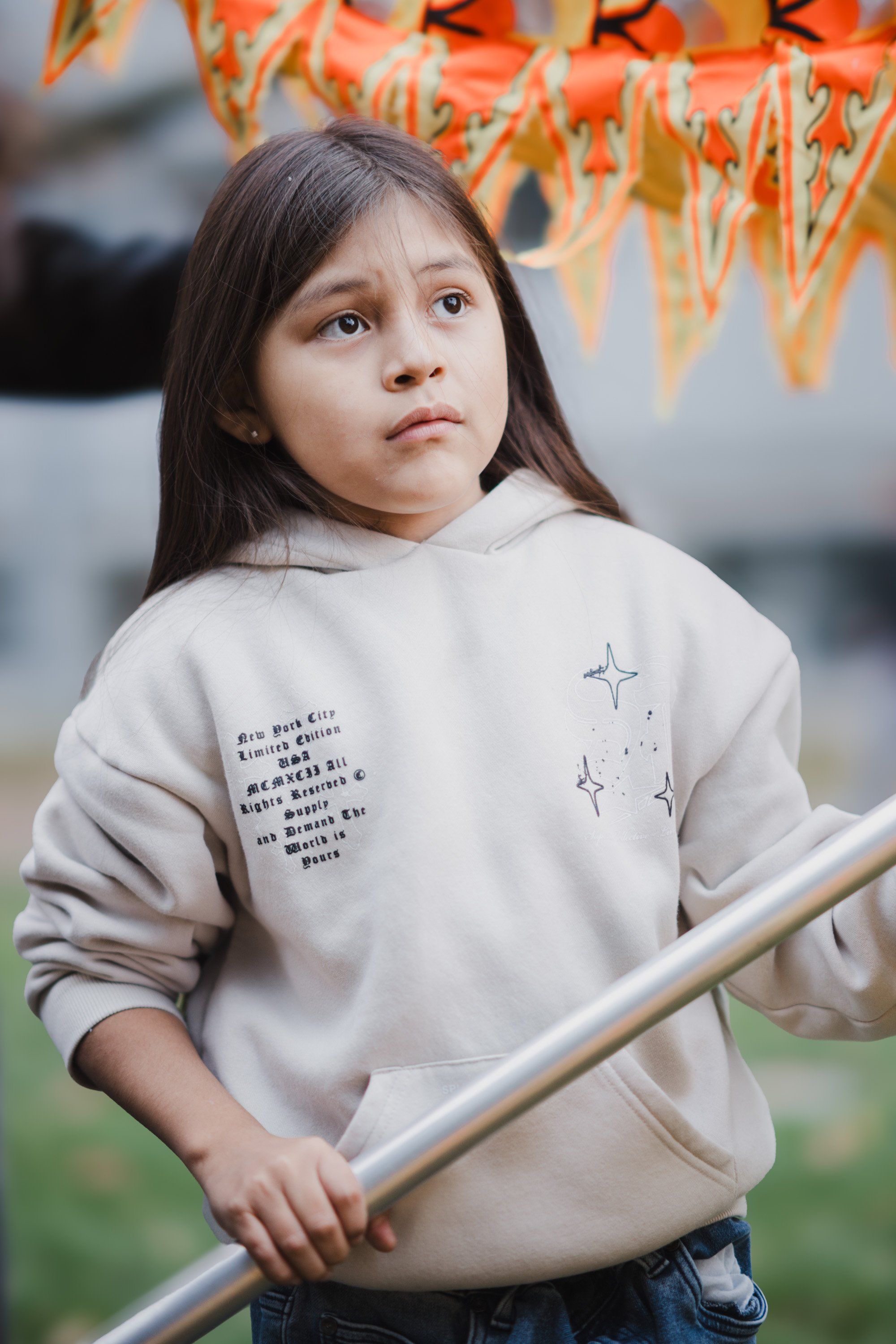 Young girl in cream hoodie with text and stars gazes upward, holding a wooden pole beneath autumn leaves