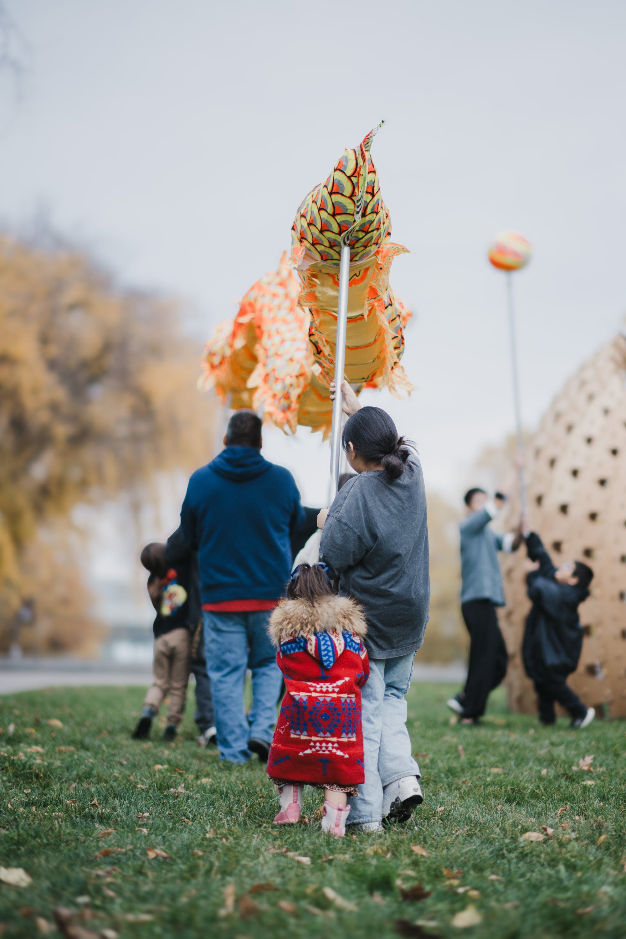 Child in red regalia held aloft holding a large cedar branch during a Coast Salish celebration, with family and community gathered on grass below