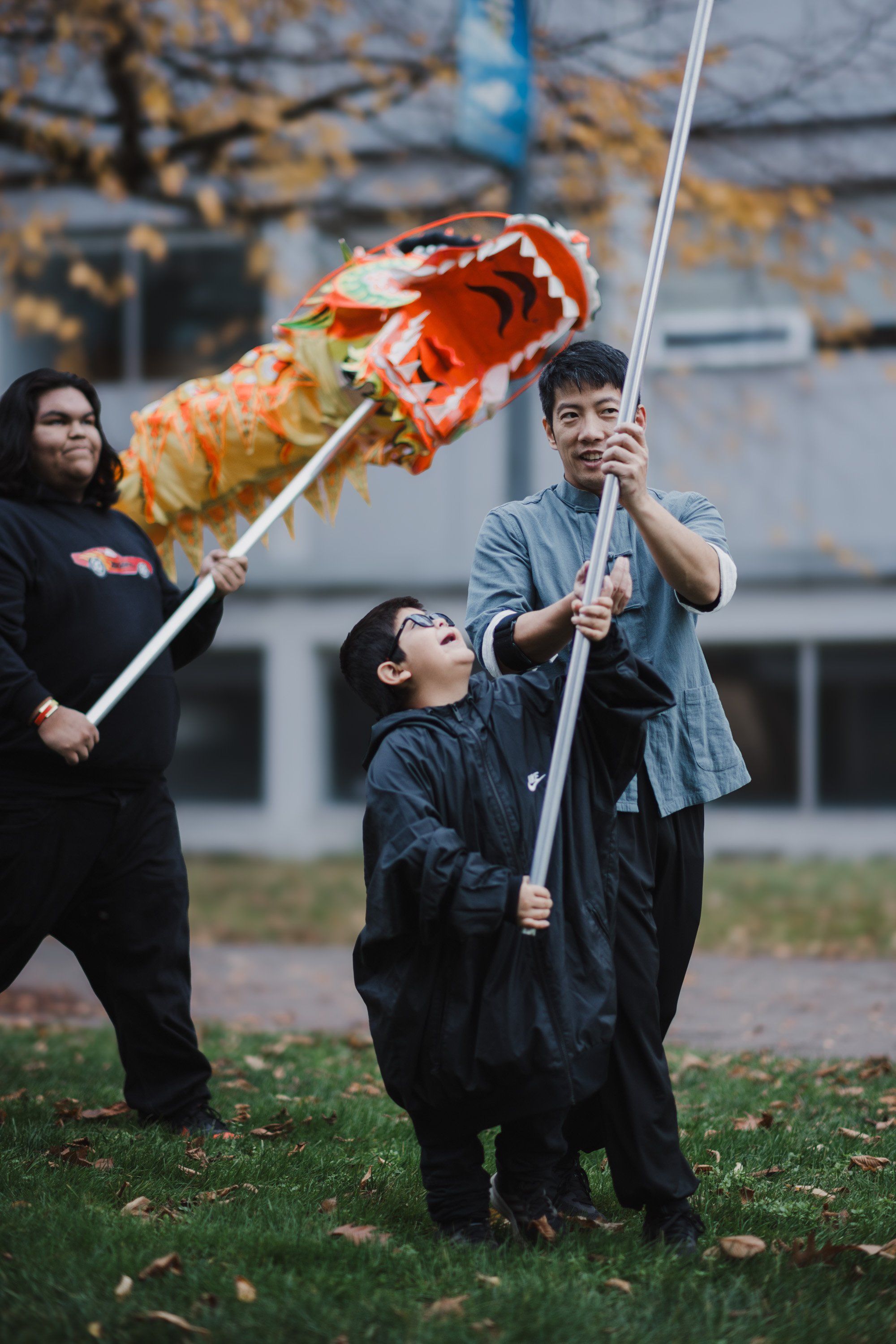 Three people hold tall poles with a colorful carved wolf puppet against a blurred building backdrop on grass