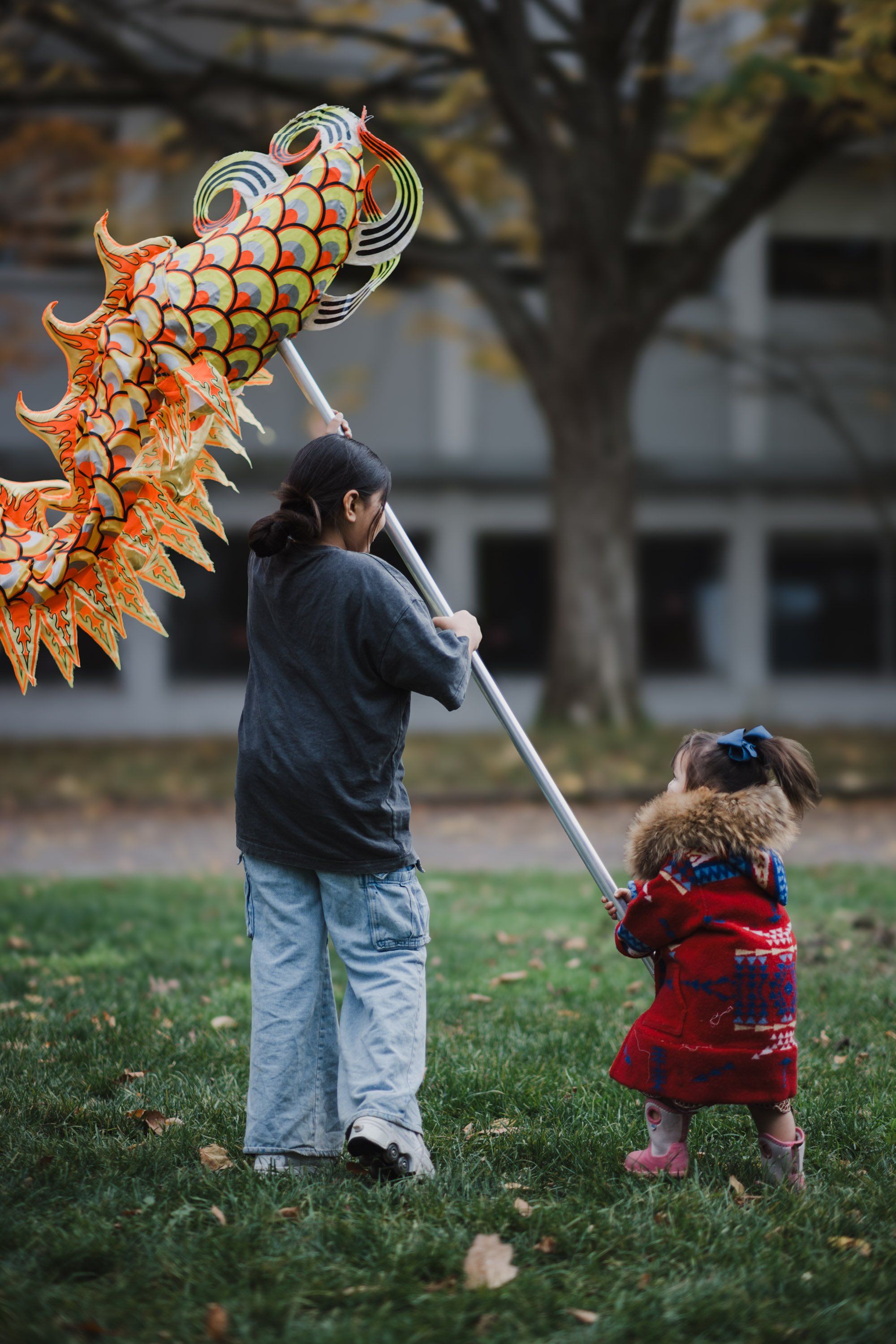 Adult and child holding a large orange decorative kite on a grass field with autumn trees and buildings in soft daylight