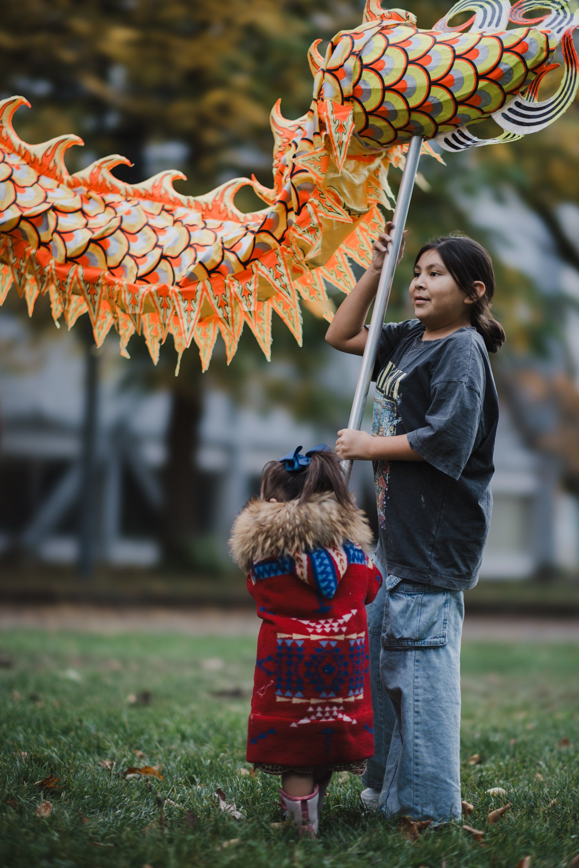 Woman and small child in red holding a large orange and white decorative dragon kite on a grass field at dusk