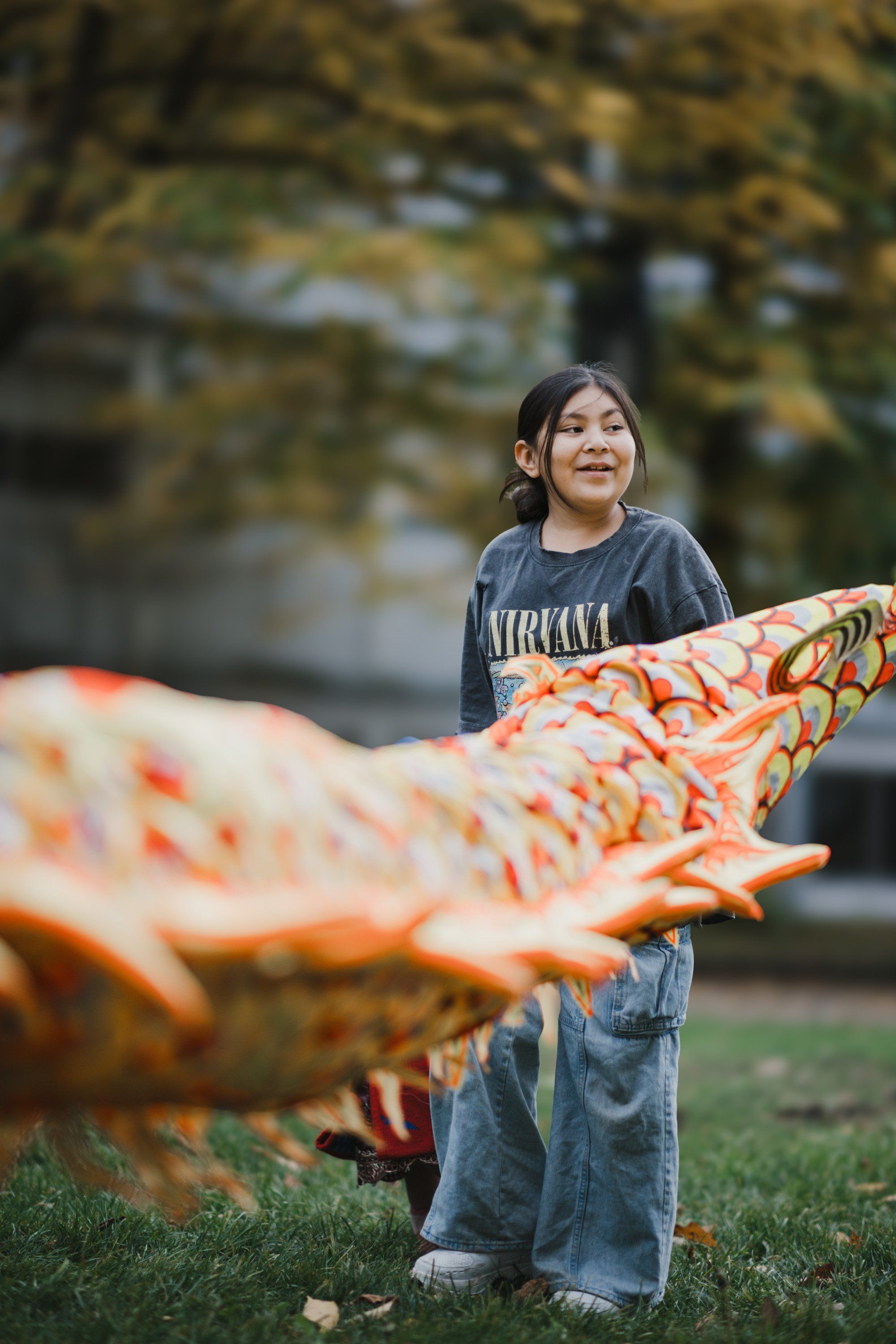Young child in Nirvana shirt holding colorful orange and red kite outdoors on grass, smiling upward with blurred evergreen trees behind
