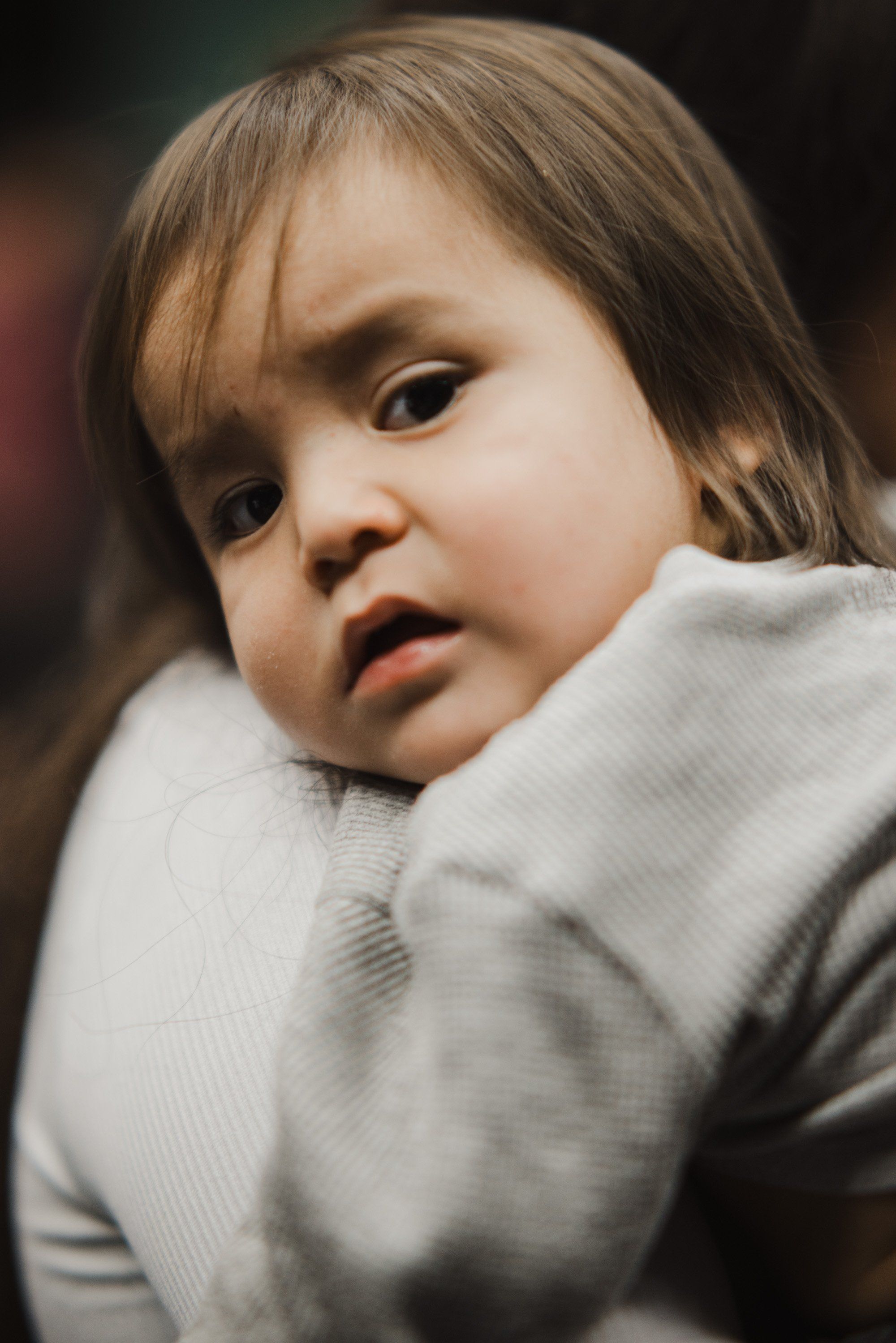 Toddler in soft gray clothing gazes thoughtfully over shoulder, warm indoor light catching gentle expression