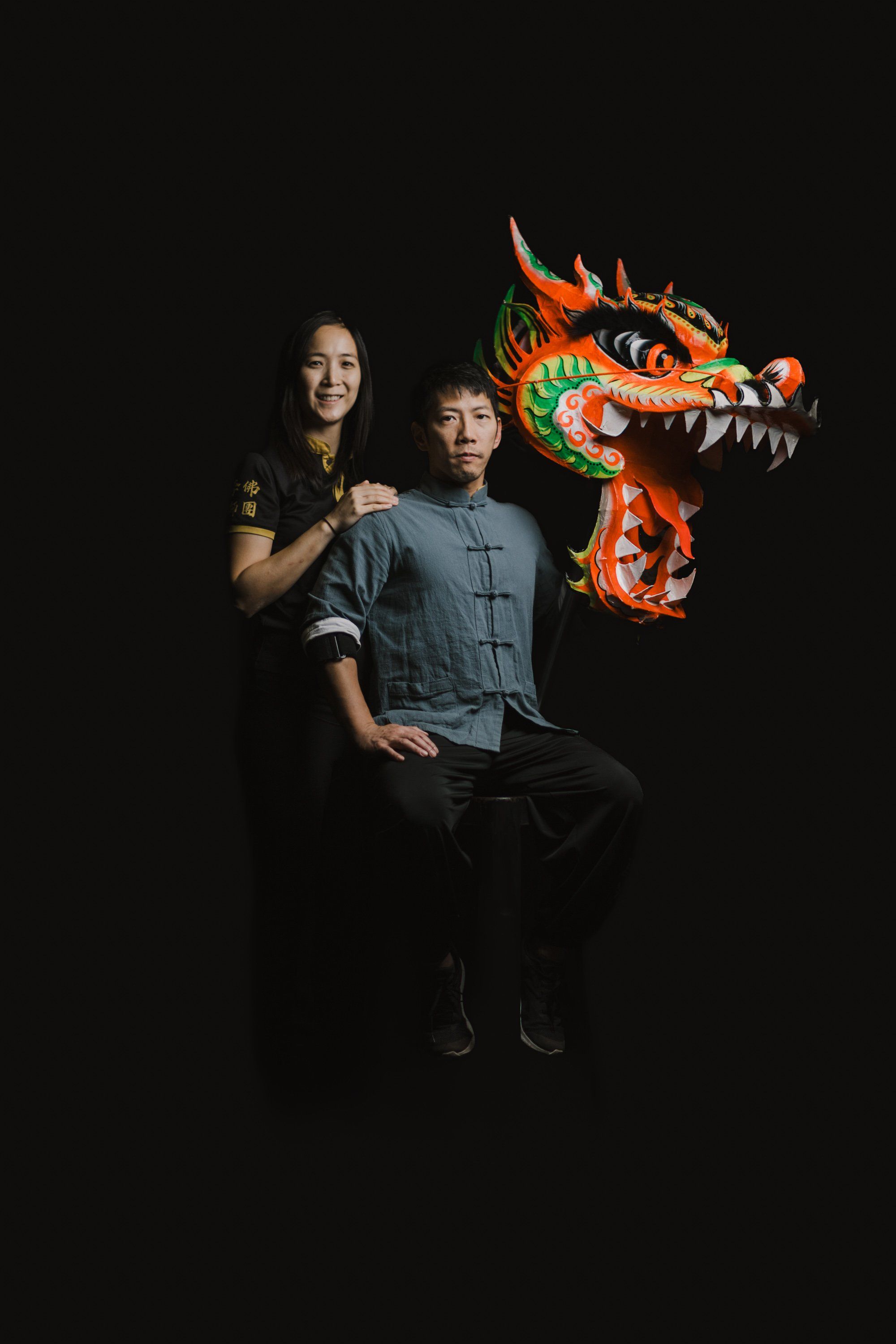 Two people pose together against a black background with a vibrant red and gold dragon figure, smiling at the camera in warm studio lighting