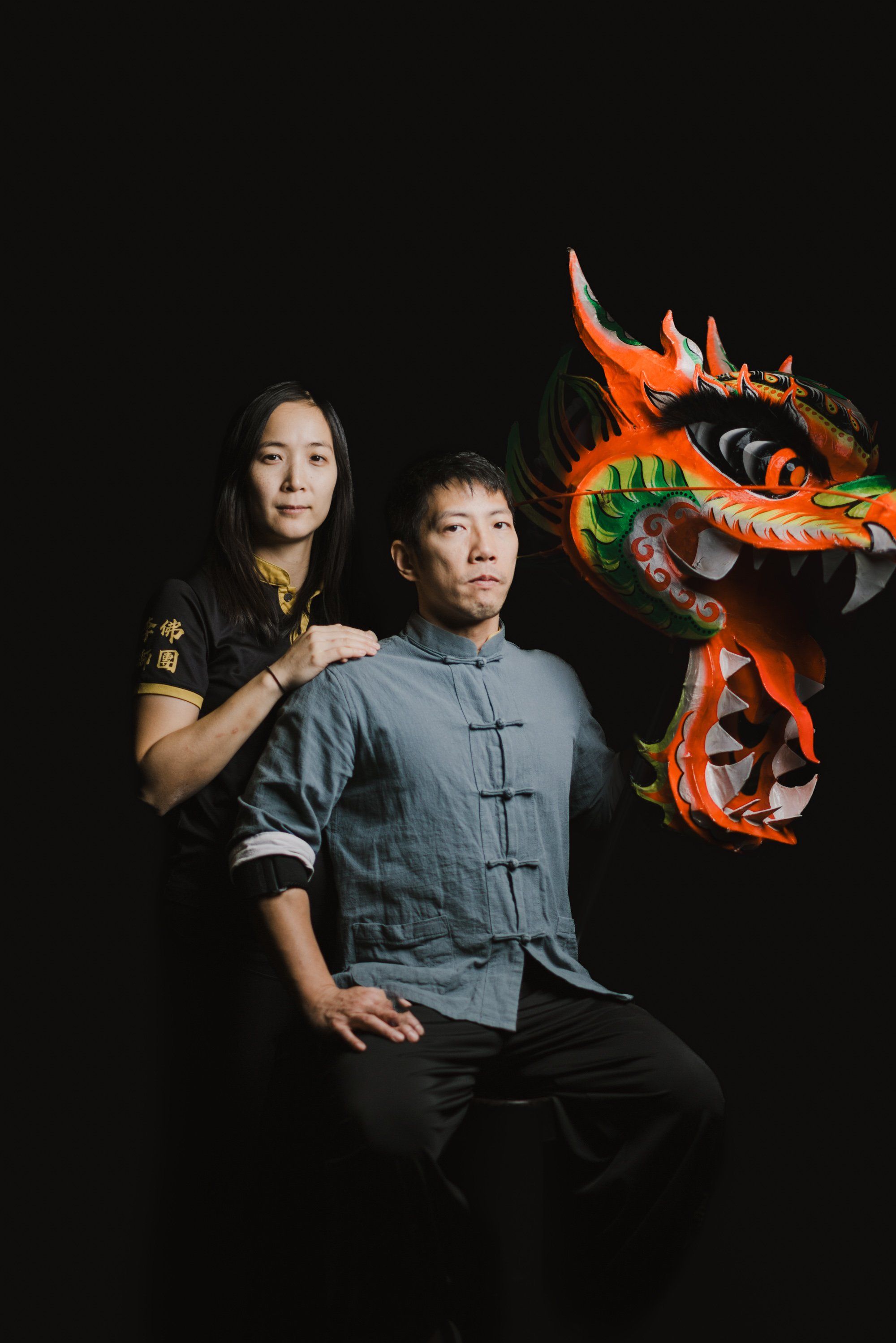 Woman in traditional regalia stands behind man in blue tunic as ornate red and gold dragon puppet looms against black background