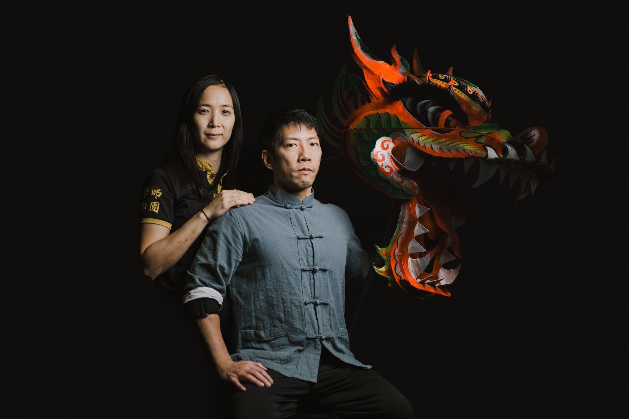 Two people pose against a black background with an ornate red and orange carved wooden dragon, dramatically lit from behind
