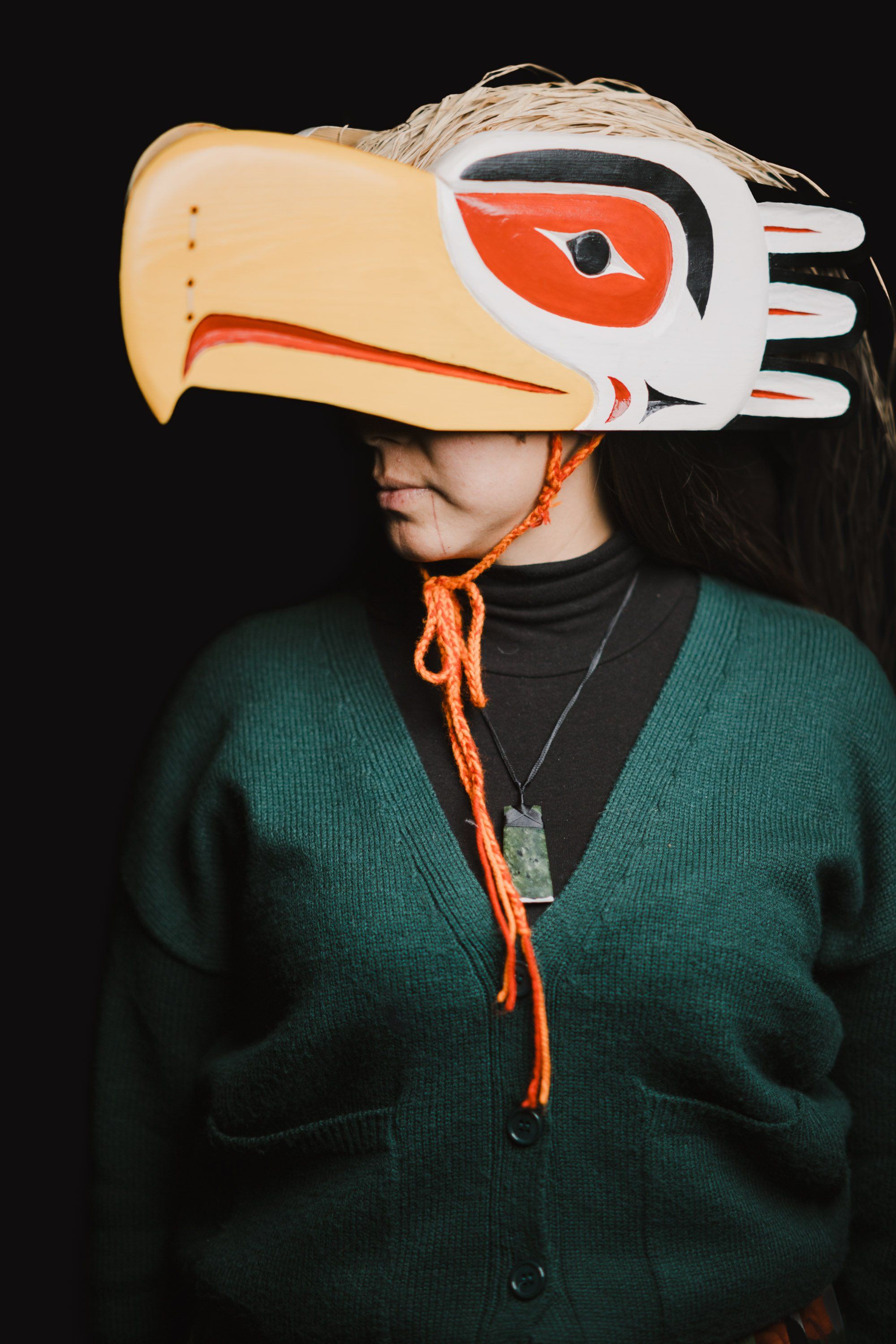 Person wearing an elaborate wooden eagle headdress with orange and white paint details, paired with a dark teal cardigan against black background