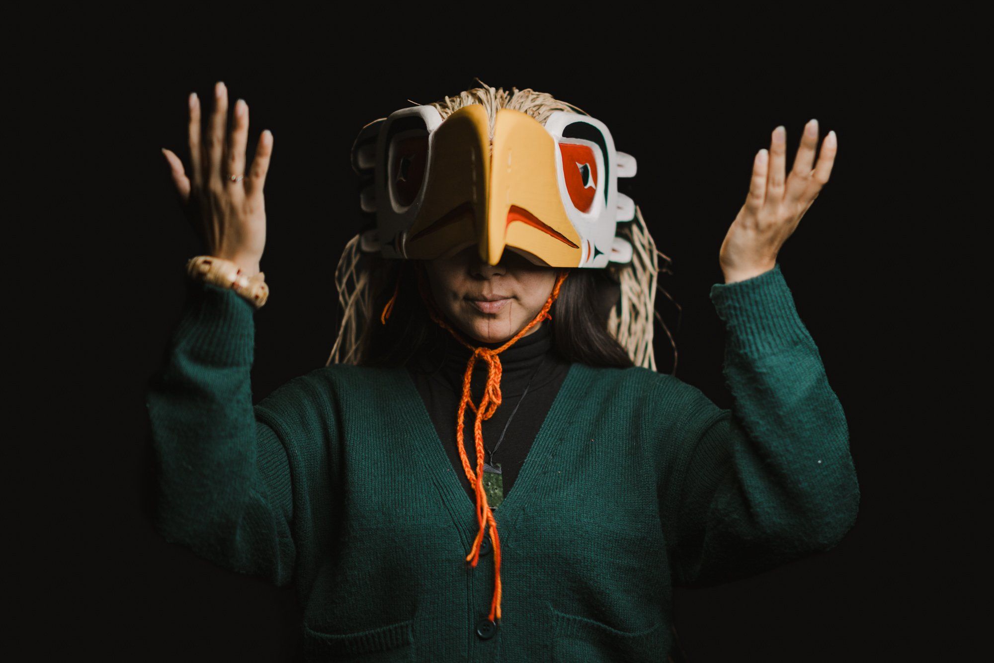 Child in traditional Coast Salish regalia with carved wooden mask and cedar hat raises hands against black background