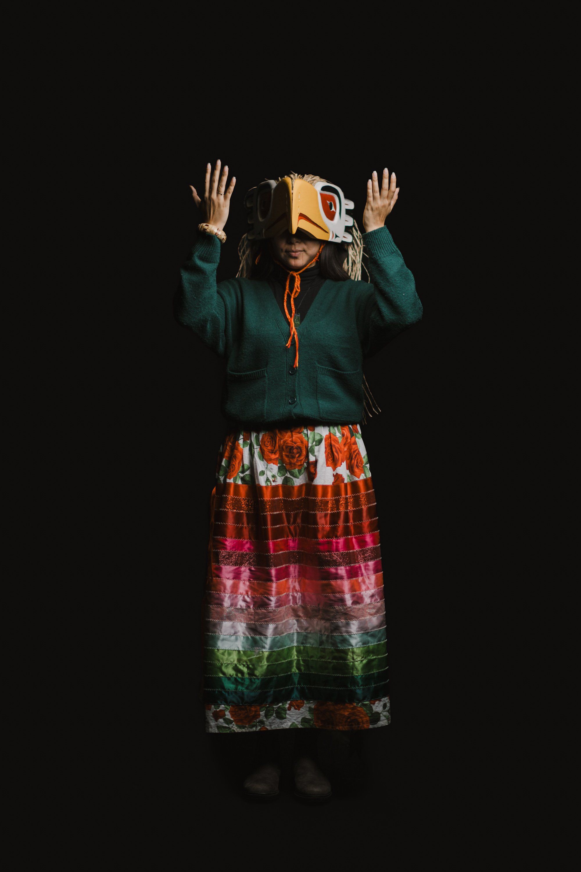 Person in forest green jacket and red plaid skirt raises hands expressively while wearing a wooden mask, against black background