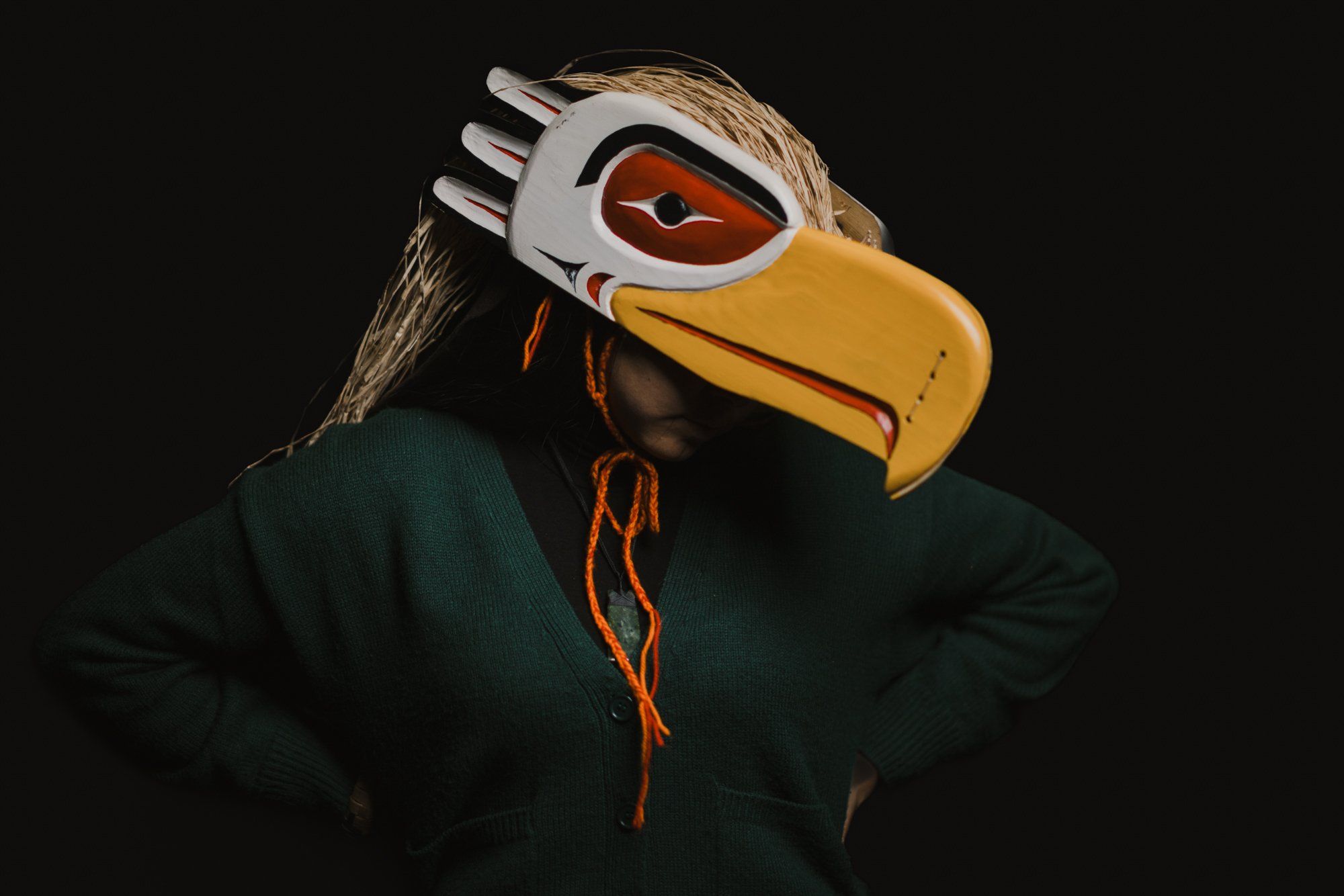 Person wearing an elaborate wooden eagle mask with white face paint and yellow beak, dark clothing against black background