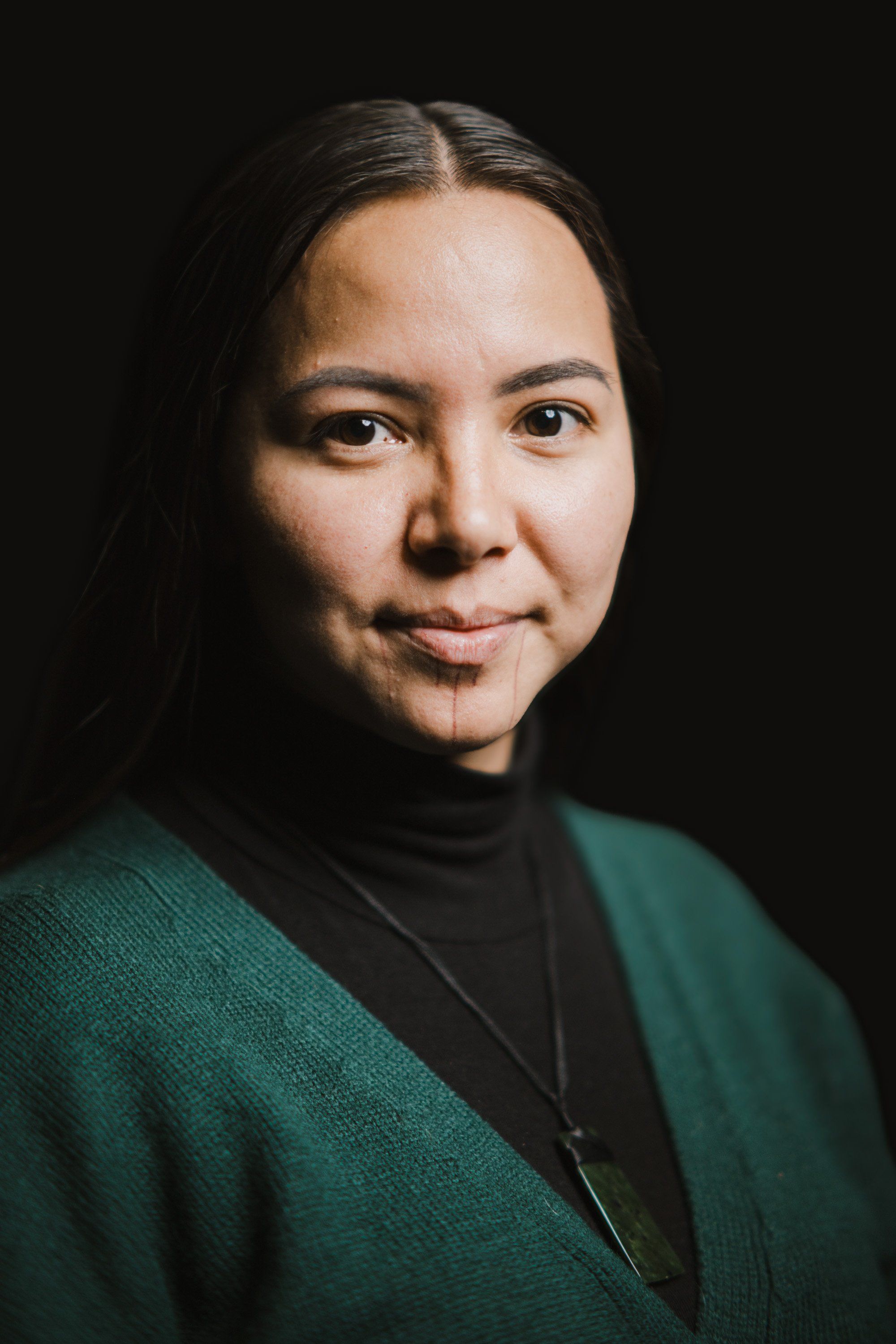 Portrait of a woman with long dark hair wearing a teal cardigan over black turtleneck against dark background, warm studio lighting