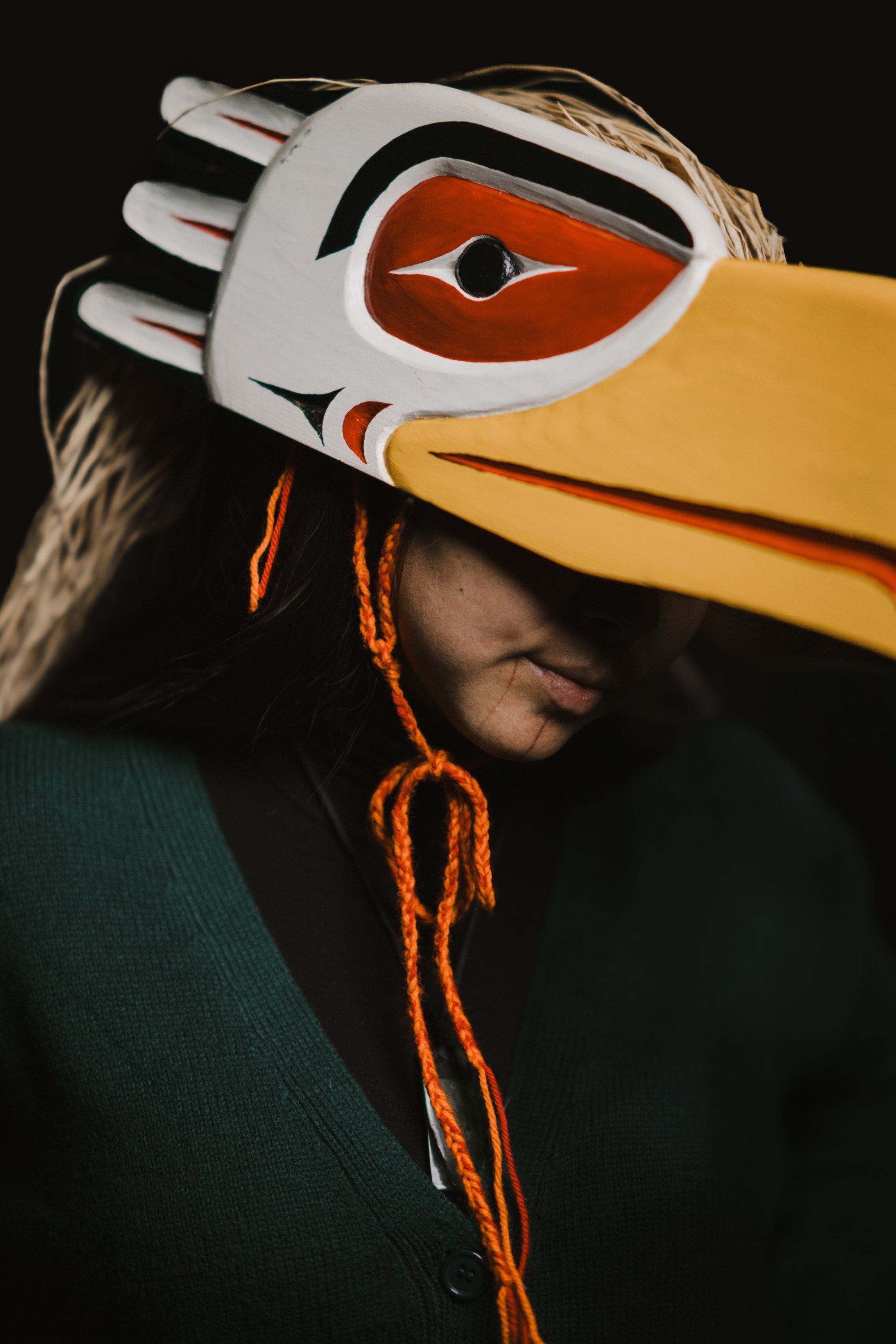 Person wearing an elaborate white and orange Coast Salish mask with dramatic eye design, holding a carved yellow cedar tool, orange cordage visible against d…