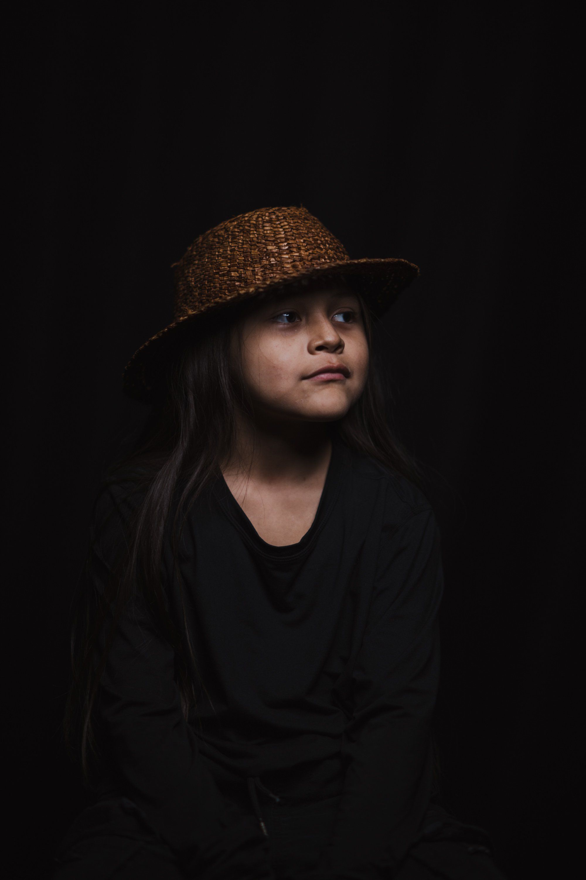 Young person in black clothing and woven hat gazes upward against dark background, warmly lit from the side