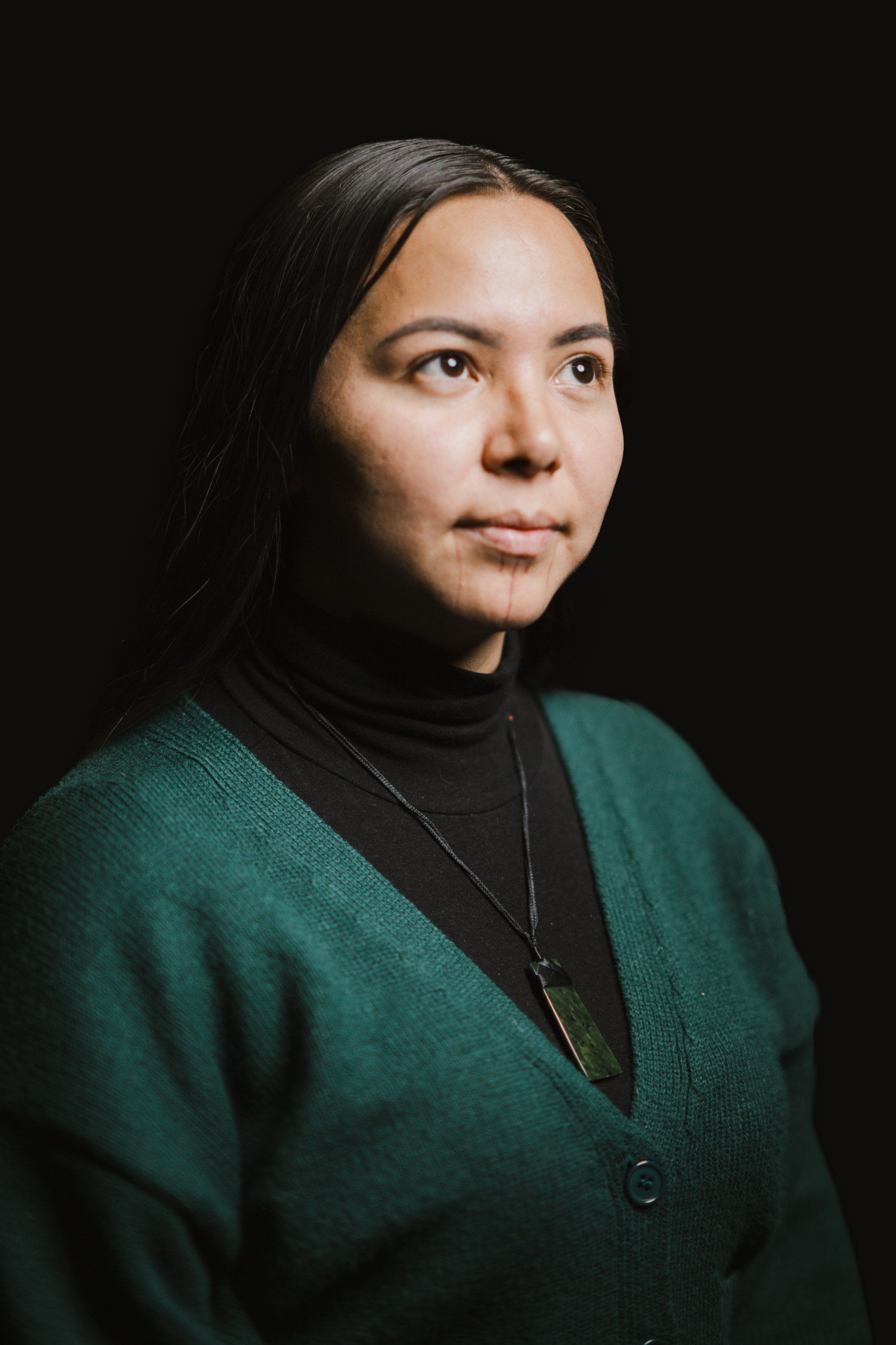 Portrait of a woman with long dark hair wearing a deep teal cardigan against a black background, looking thoughtfully to the side