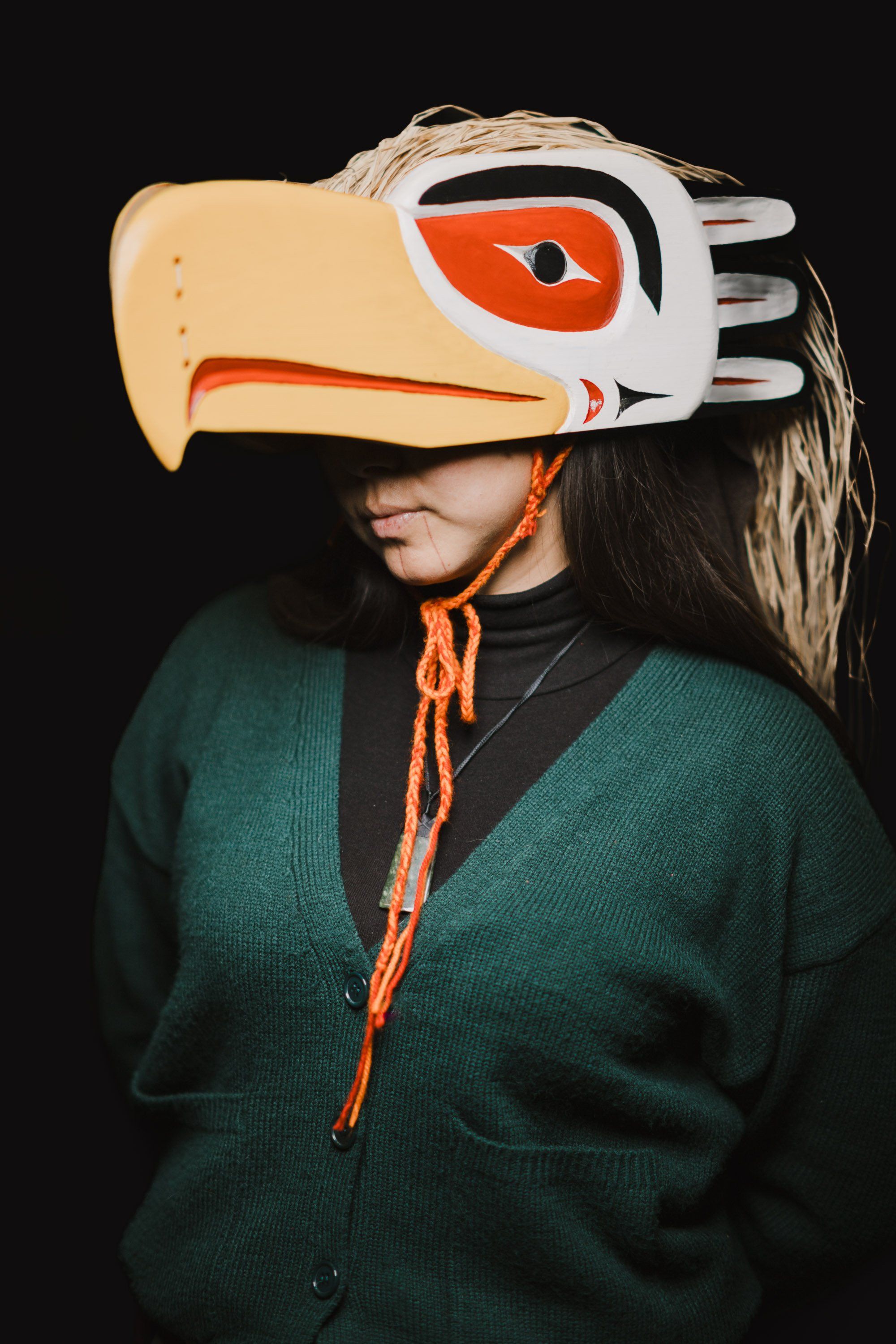 Person wearing an oversized carved wooden eagle mask in white, orange and black, with long dark hair visible, dressed in a teal cardigan against a black back…