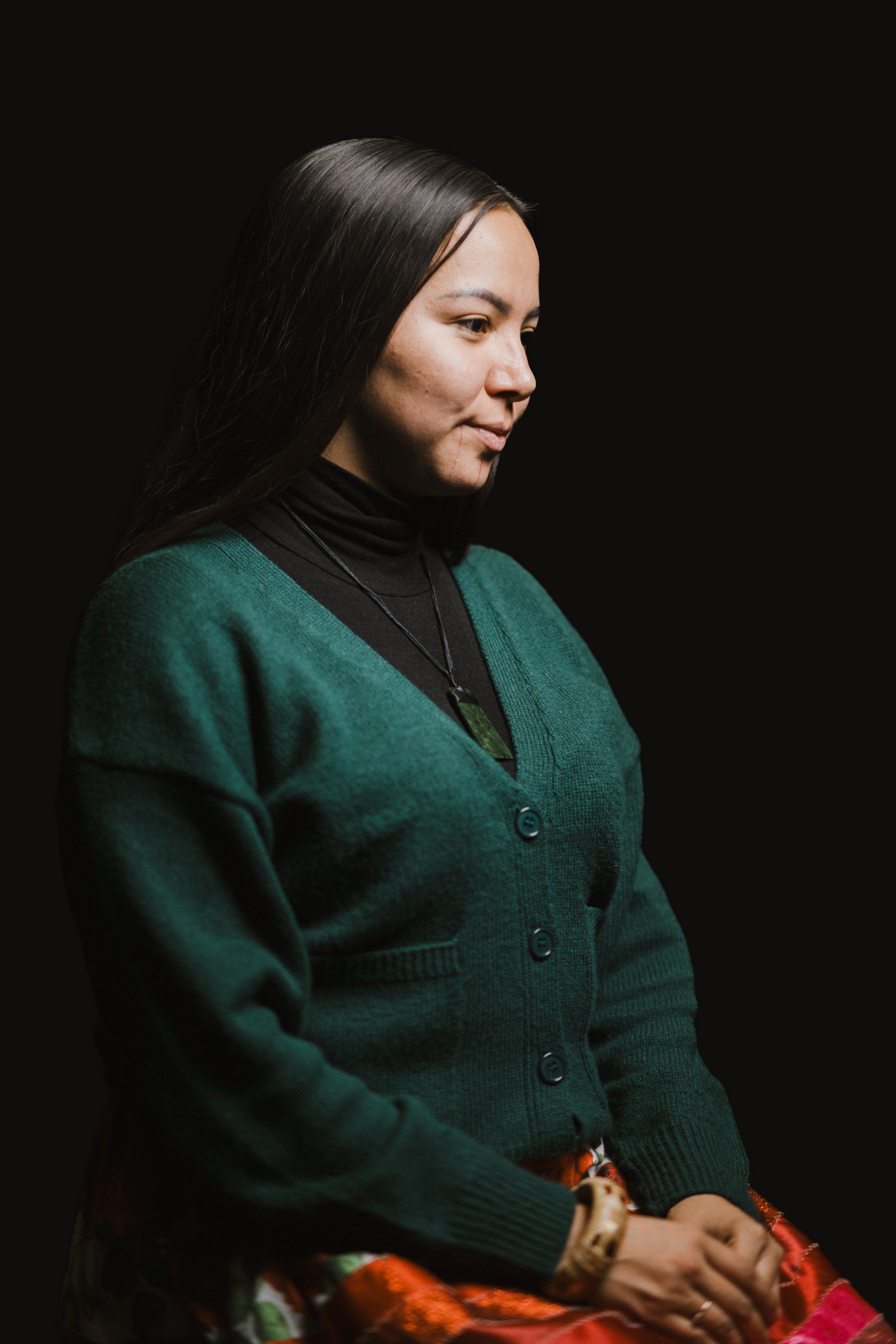Woman in forest-green cardigan and black turtleneck gazes thoughtfully leftward against black background, hands folded with colorful fabric visible below