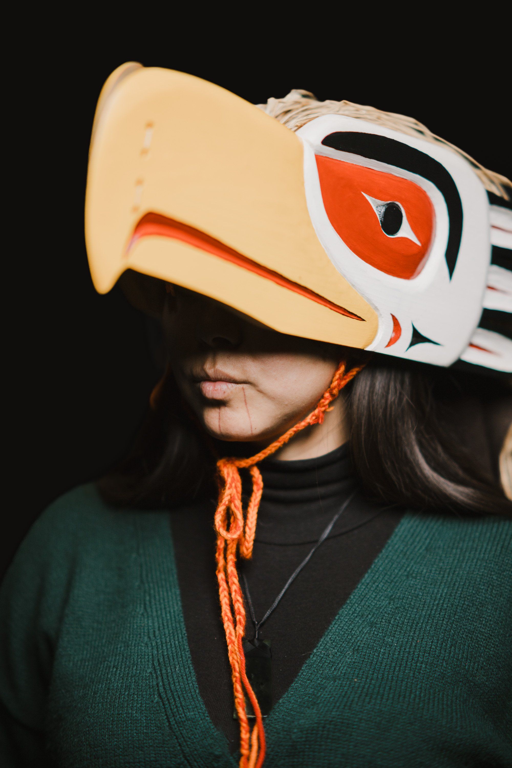 Person wearing an elaborate cream and orange Coast Salish mask with geometric designs, dark long hair visible, against a black background