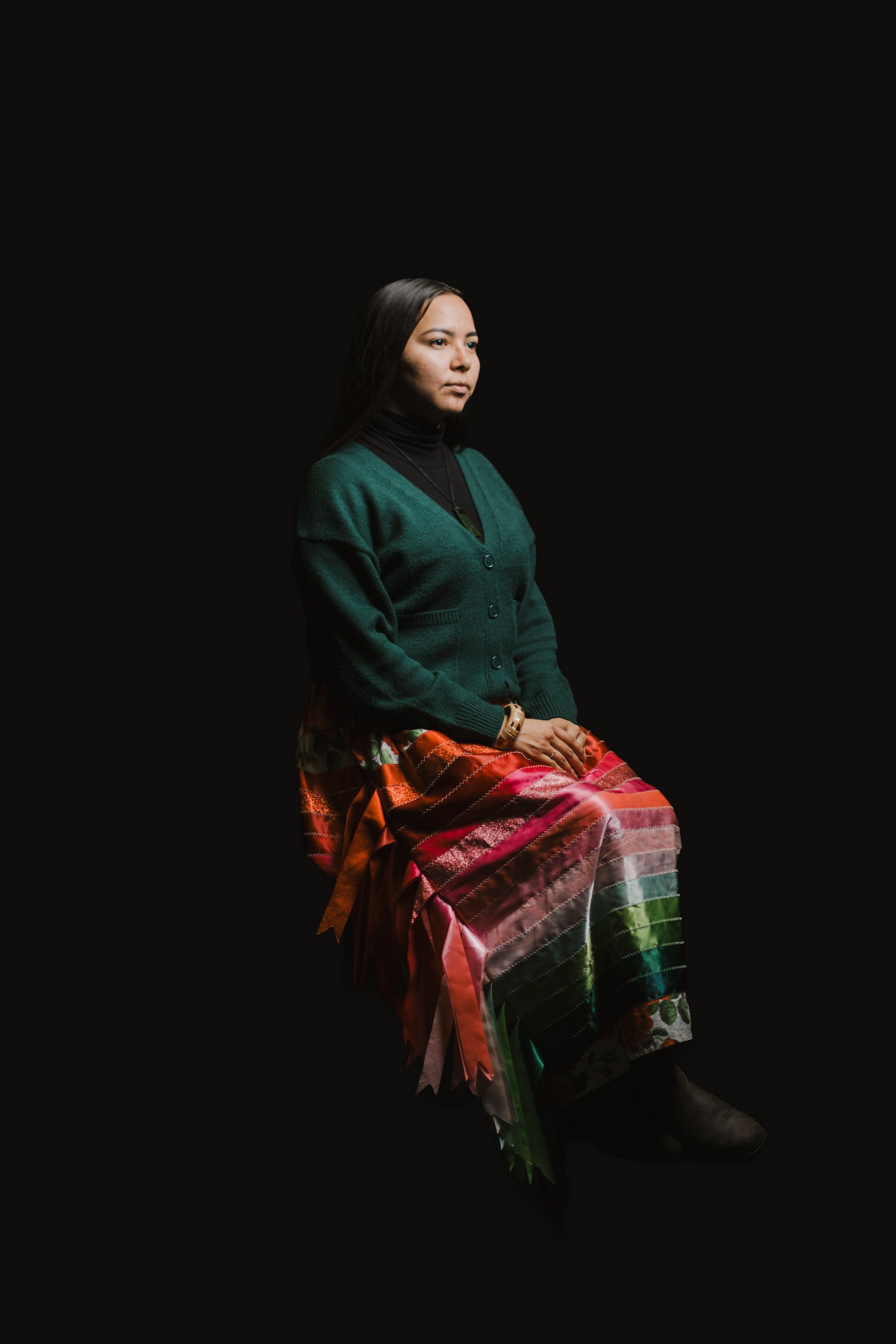 Woman in dark green cardigan and vibrant red plaid shawl seated against black background, gazing thoughtfully to the side