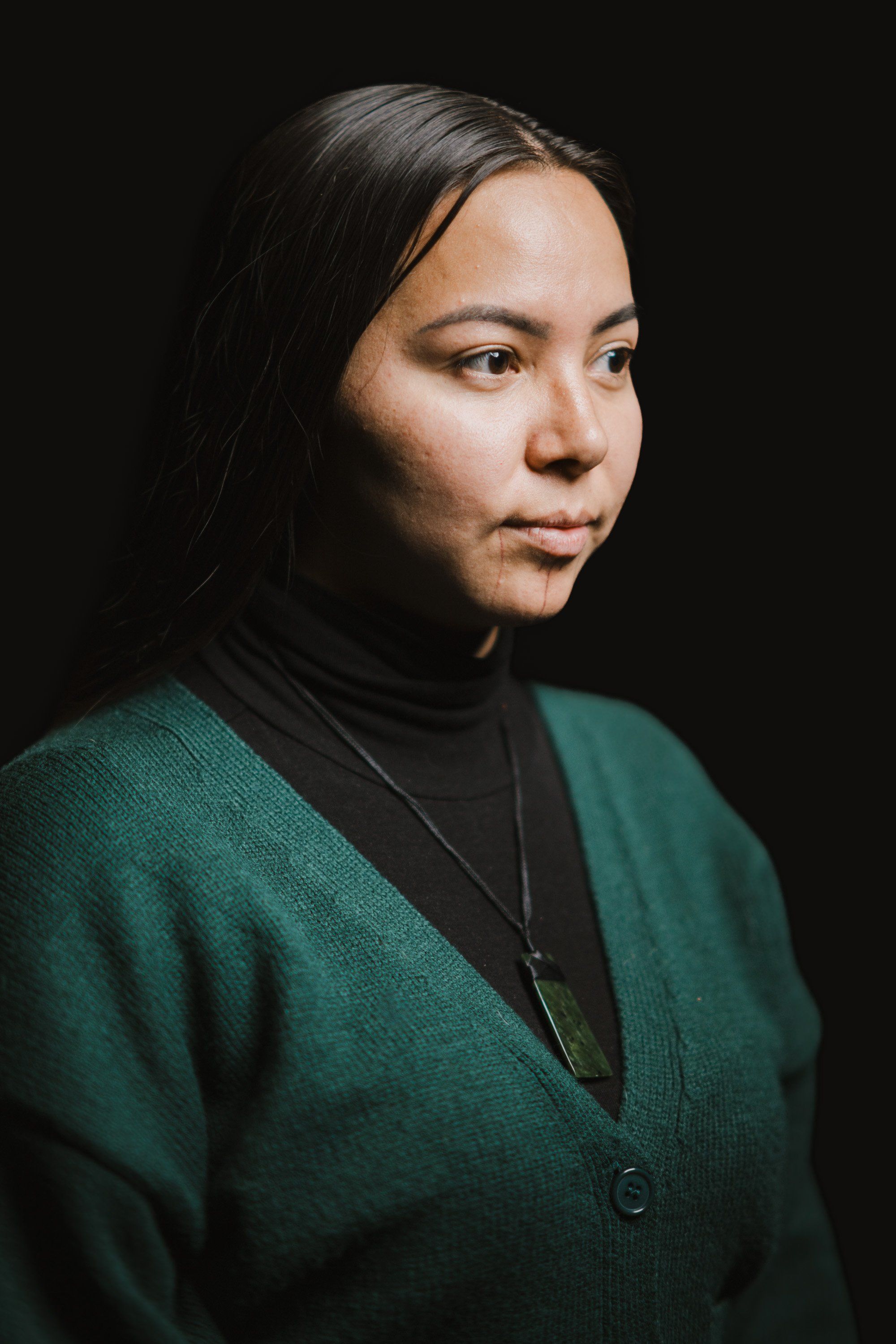 Woman in forest green cardigan and black turtleneck gazes thoughtfully to the side against dark background