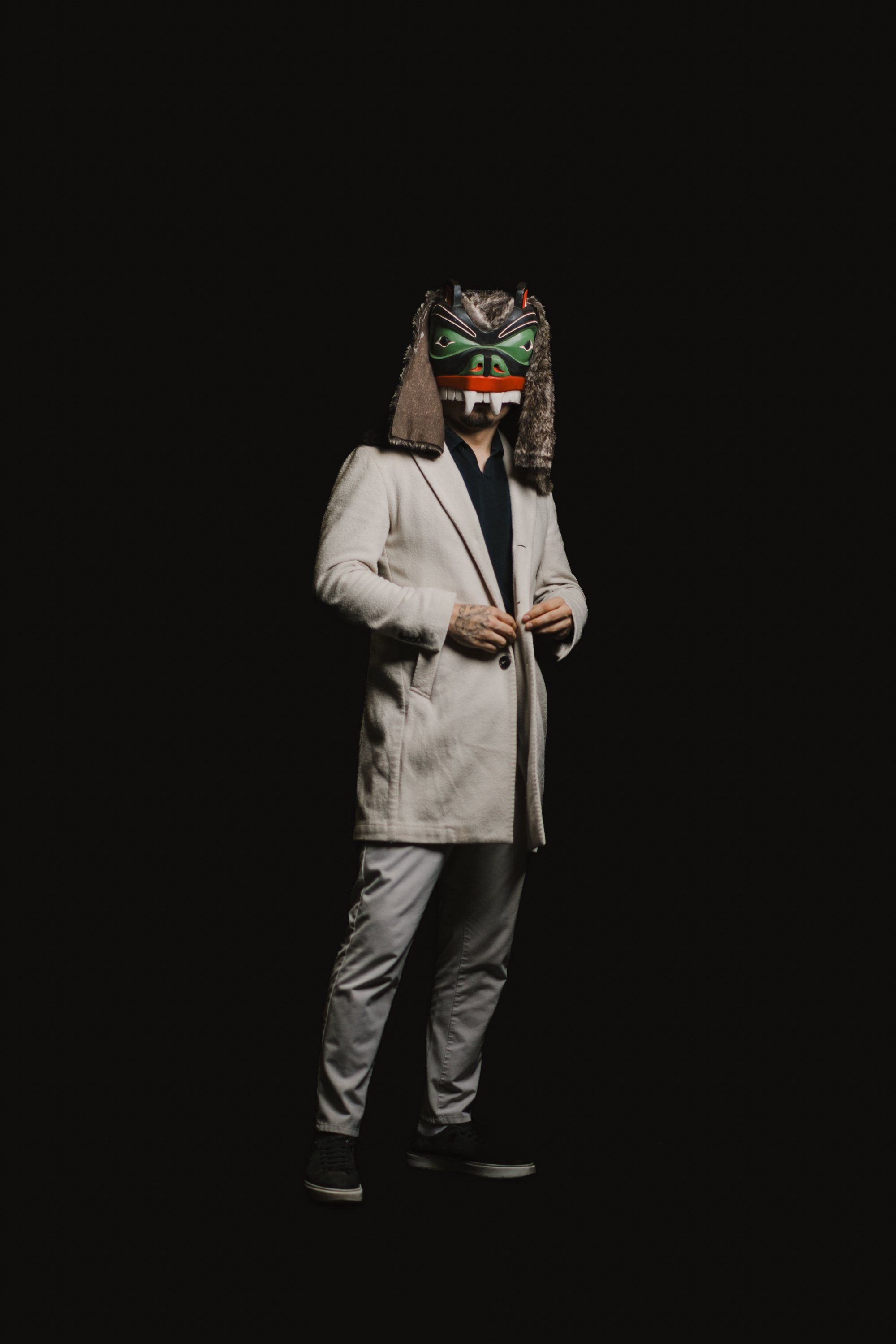 Person in light grey blazer and dark pants stands against black background, wearing an ornate carved mask with green and copper tones and long hair attachment