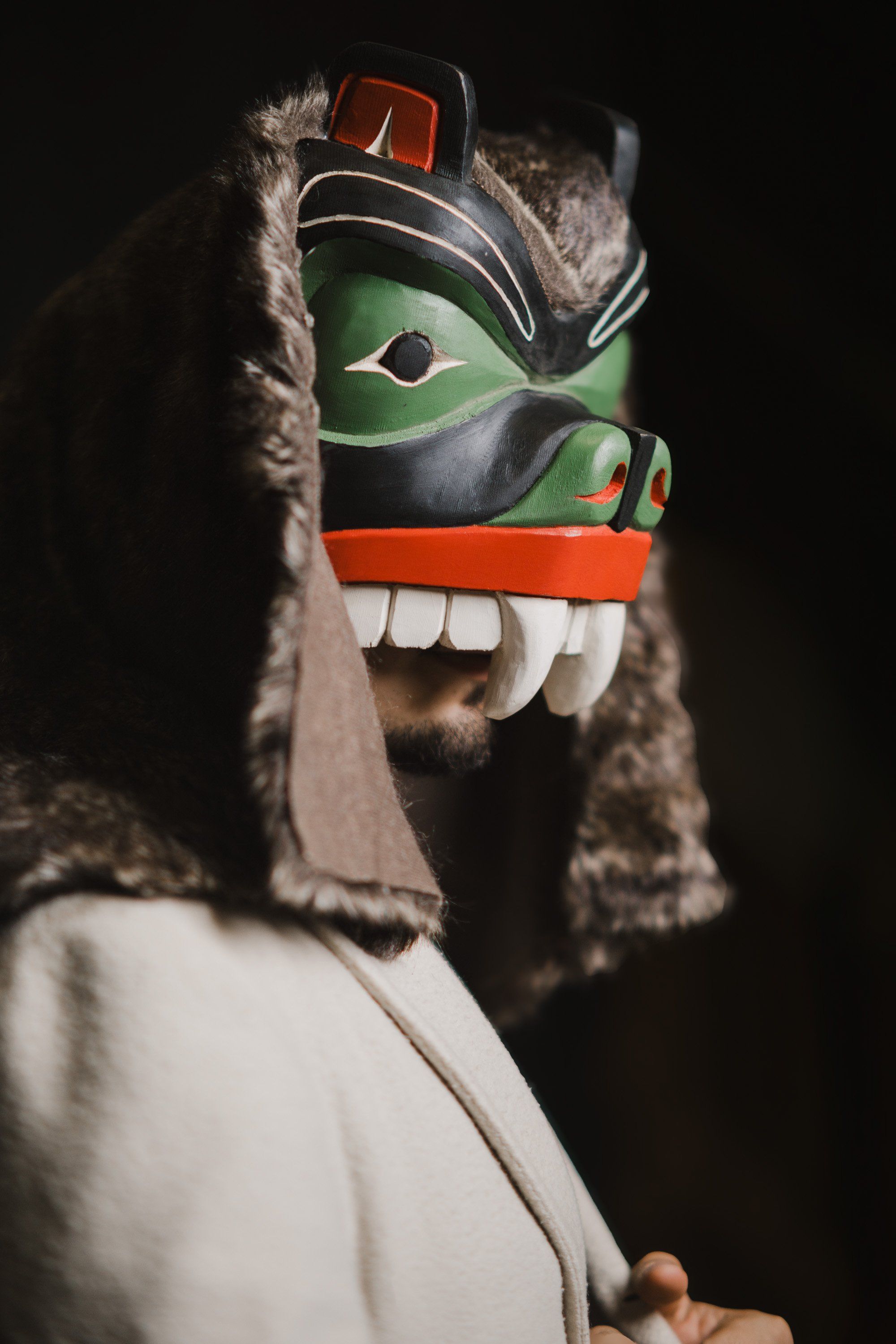 Person wearing an elaborate Coast Salish wolf mask with green face paint, white teeth, and fur trim, photographed against black background