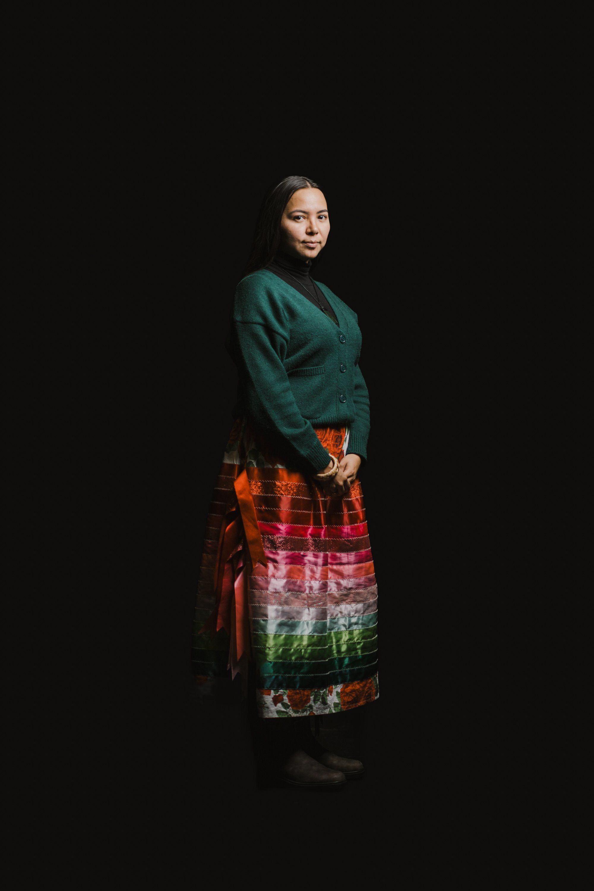 Woman in teal sweater and vibrant plaid skirt with traditional patterns stands against black background, turning toward camera with gentle expression