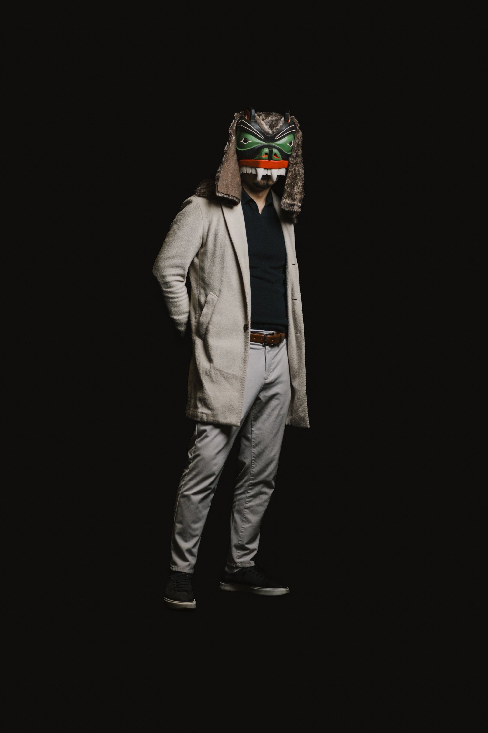 Person in light-colored suit and long cardigan wearing an elaborate green-eyed wolf mask, posed against black background