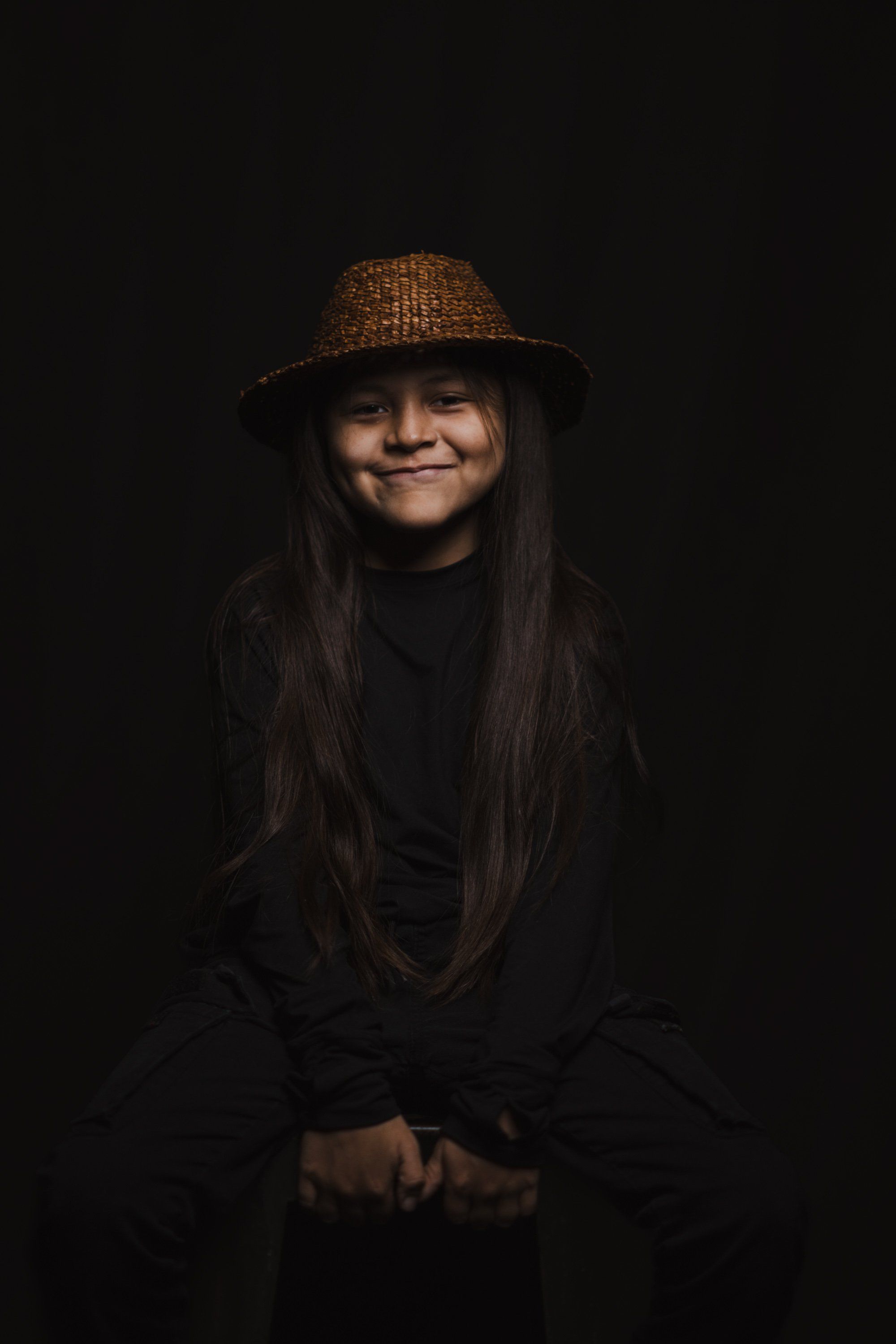 Portrait of a smiling person wearing a brown woven hat and black clothing against a dark background, warmly lit from above