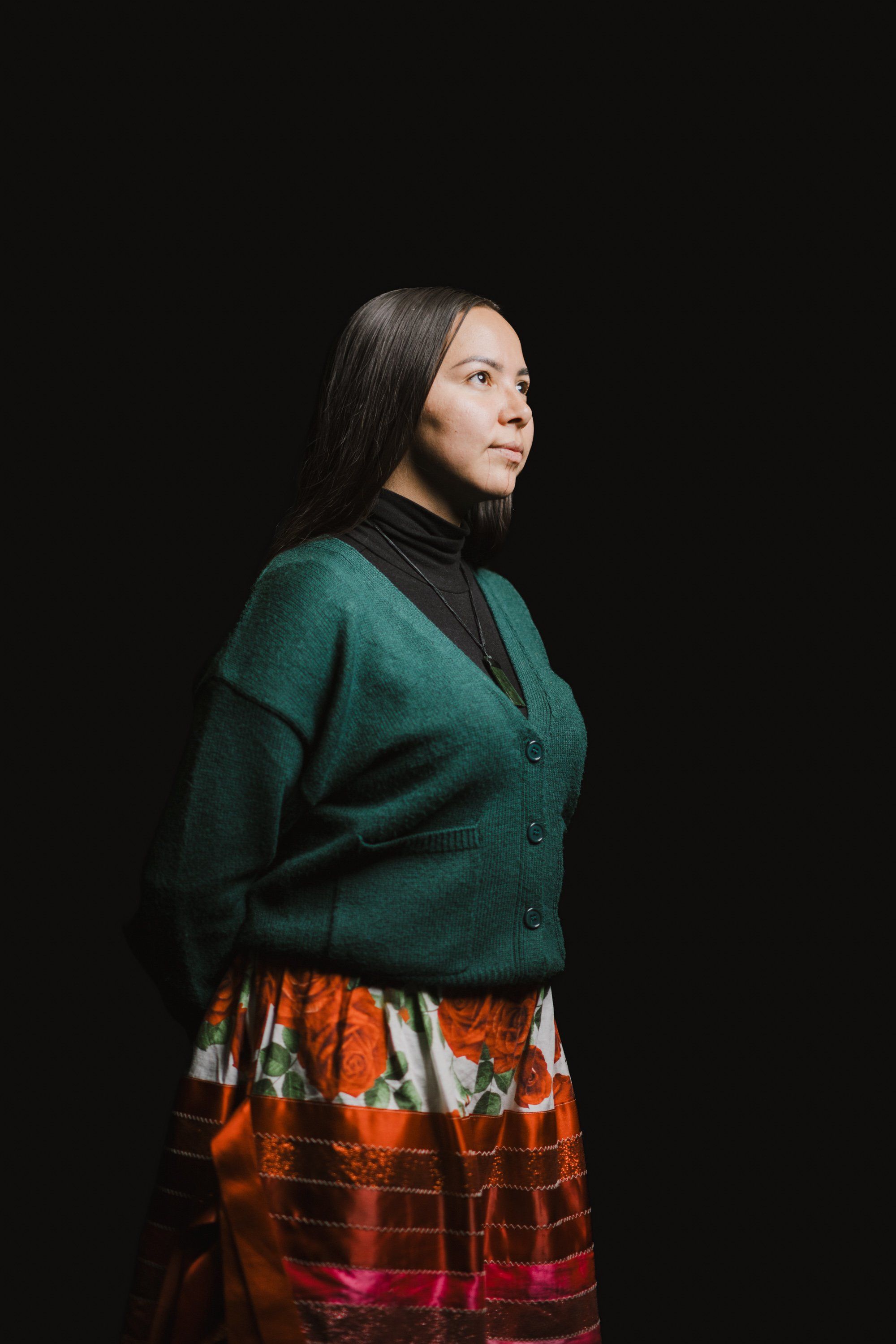 Woman in forest-green cardigan and orange plaid skirt gazes thoughtfully to the side against black background
