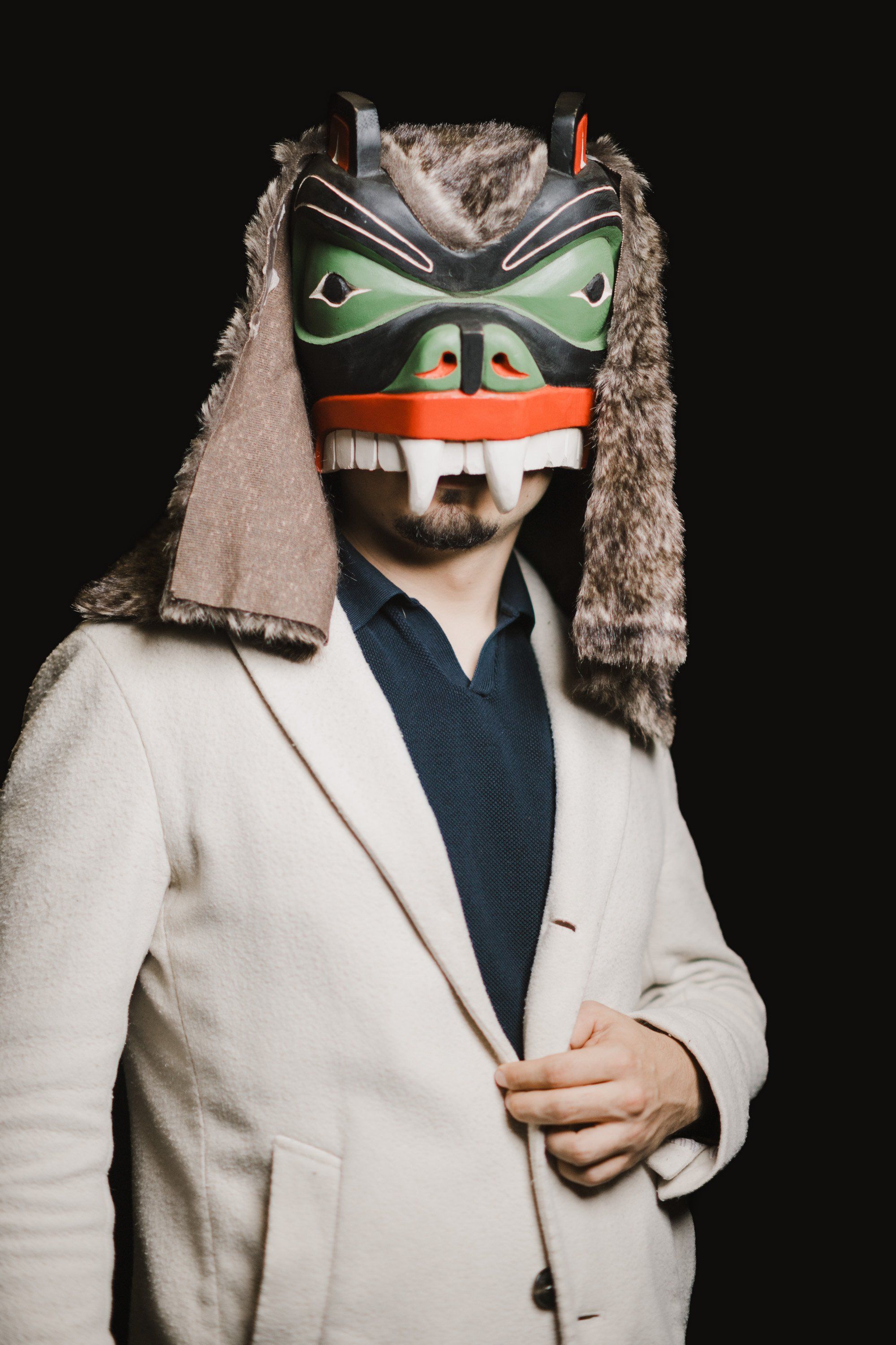 Man in cream blazer wearing an elaborate Coast Salish mask with green eye designs and fur regalia against black background