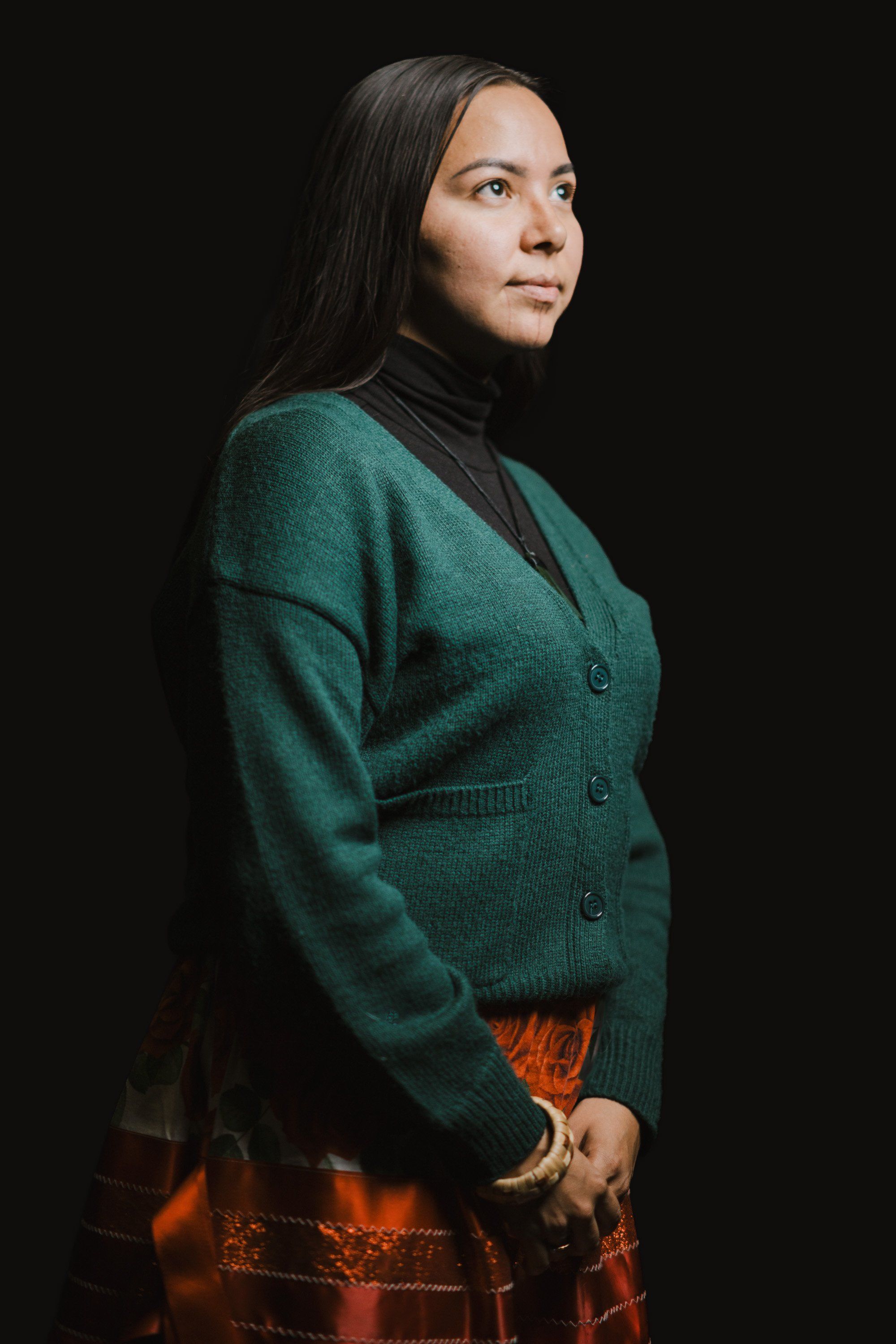 Woman in teal cardigan and orange plaid skirt gazes thoughtfully over her shoulder against dark background