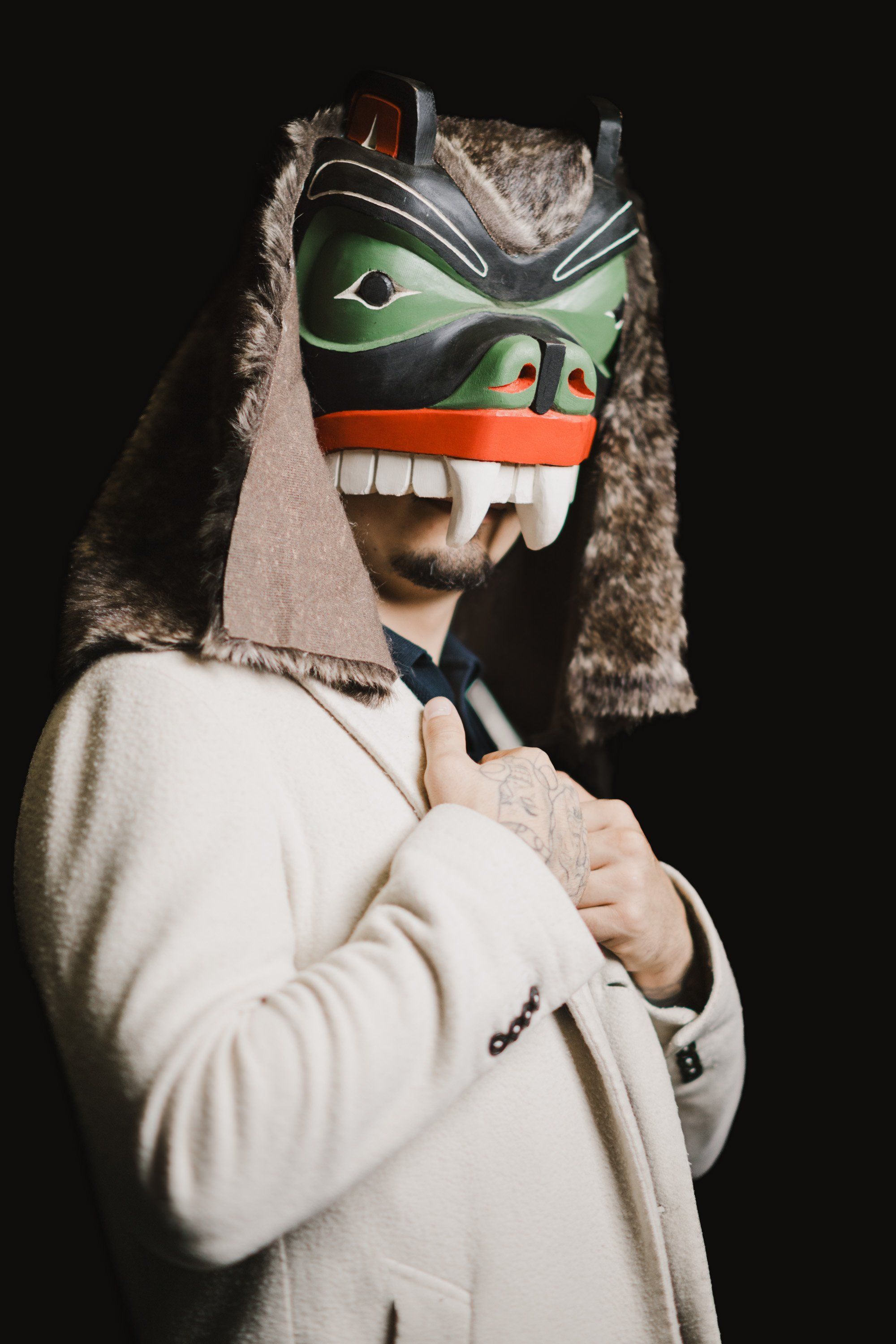 Person in cream blazer wearing traditional Coast Salish mask with green, black and orange face paint, fur-lined hood, against black background
