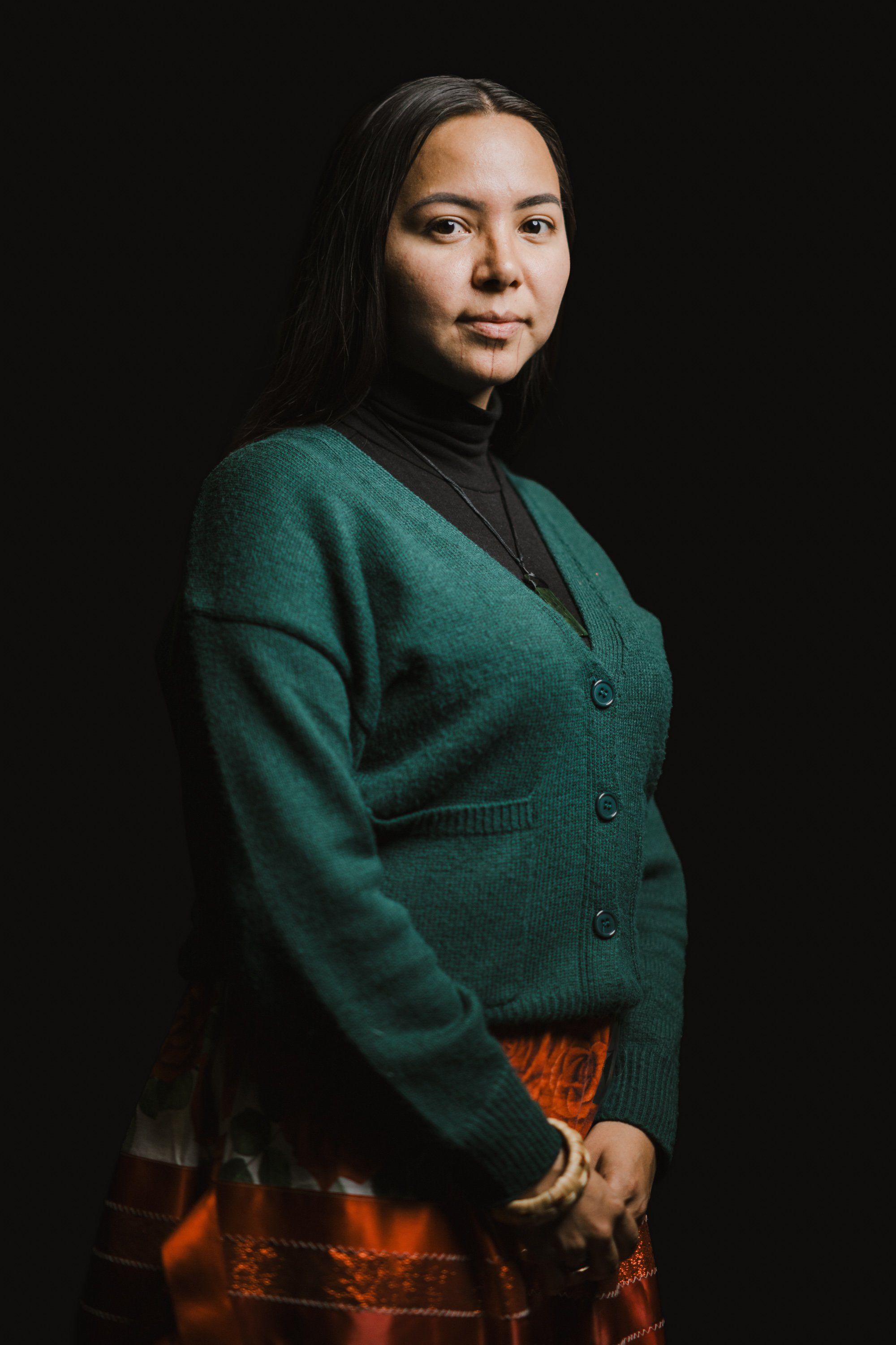 Woman in forest-green cardigan and plaid skirt gazes over shoulder against dark background, warm studio lighting