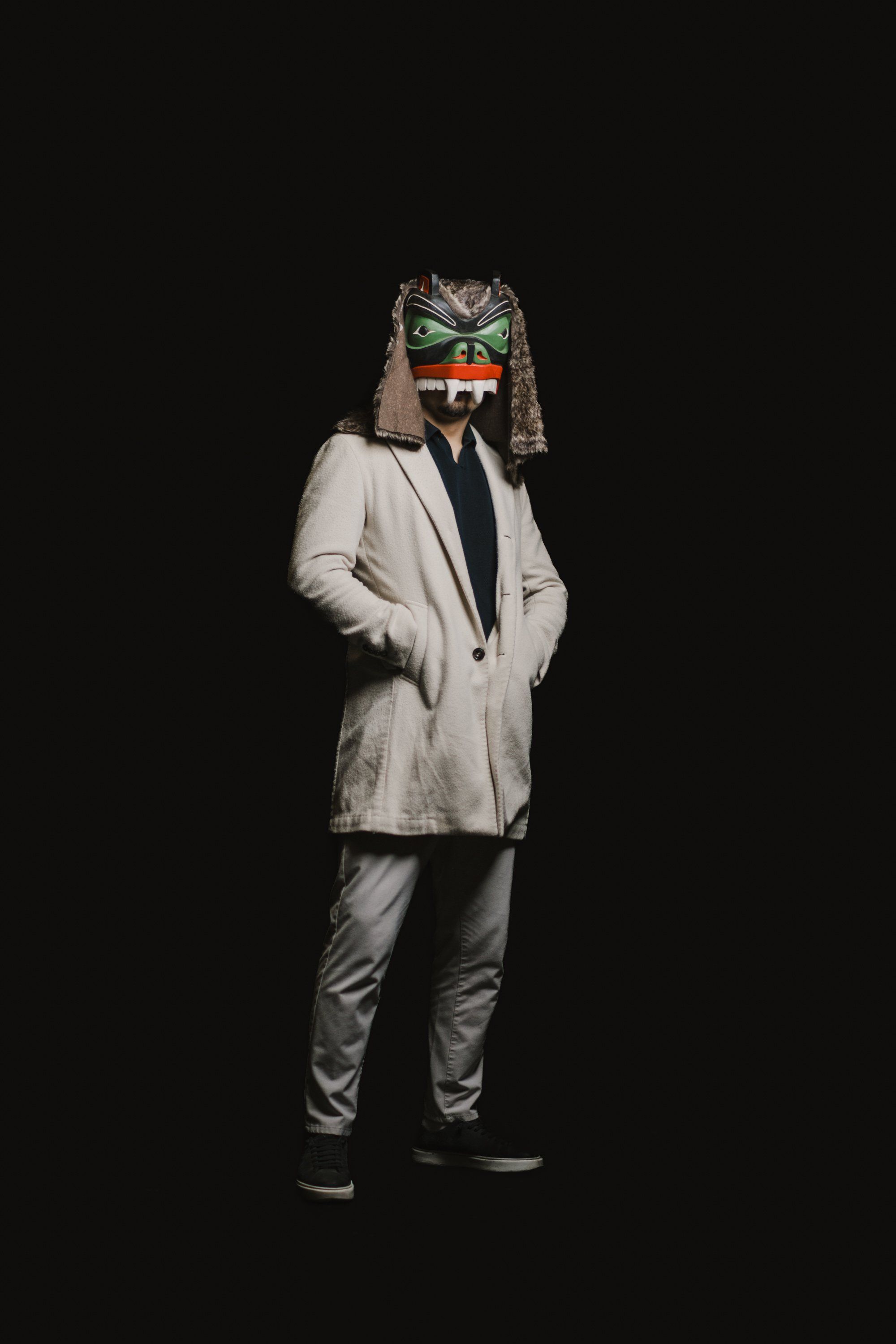 Person in light blazer with carved wolf mask and traditional regalia stands against black background, hands in pockets